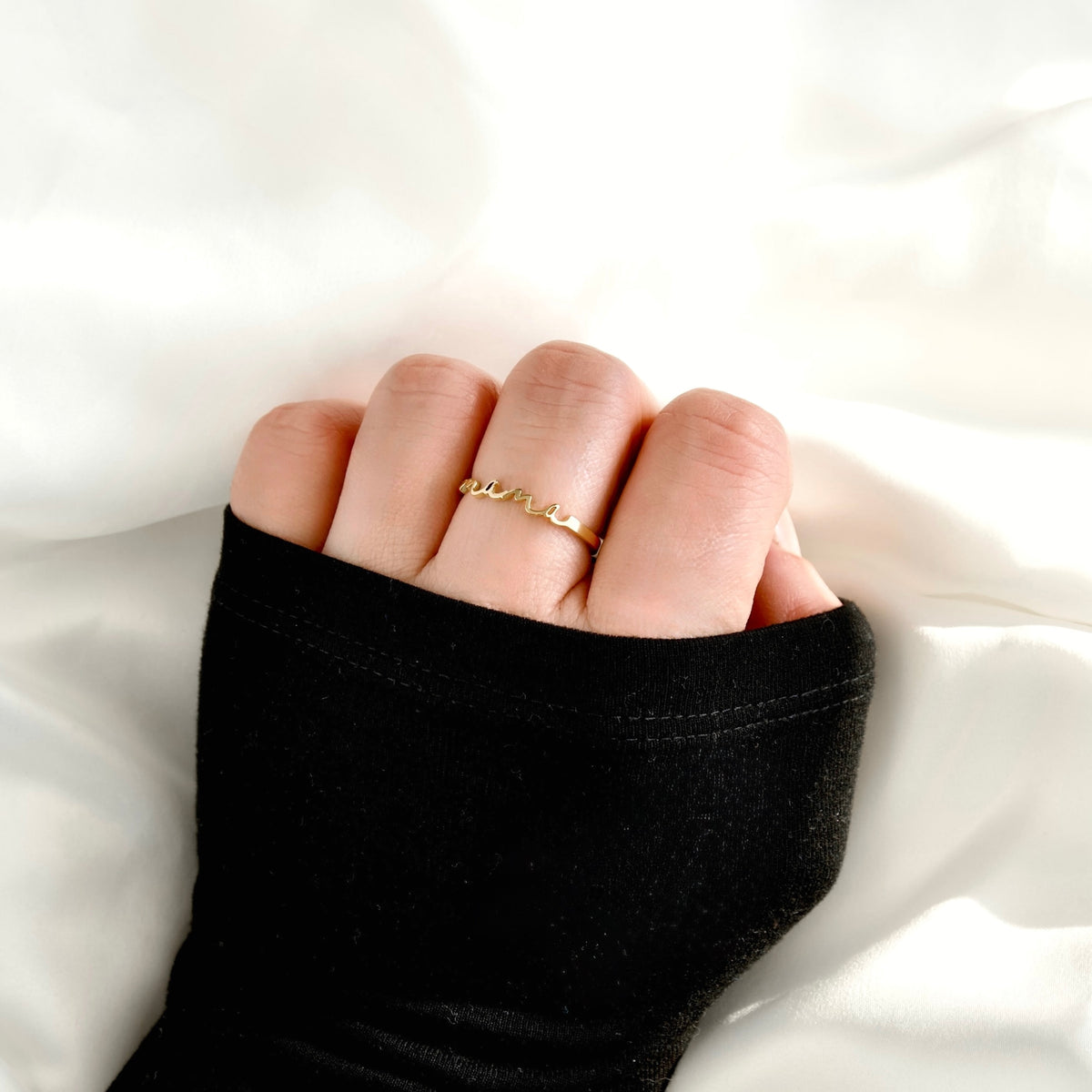 A close-up of a hand wearing a delicate gold mama ring, covered by a black sleeve, resting on white fabric.