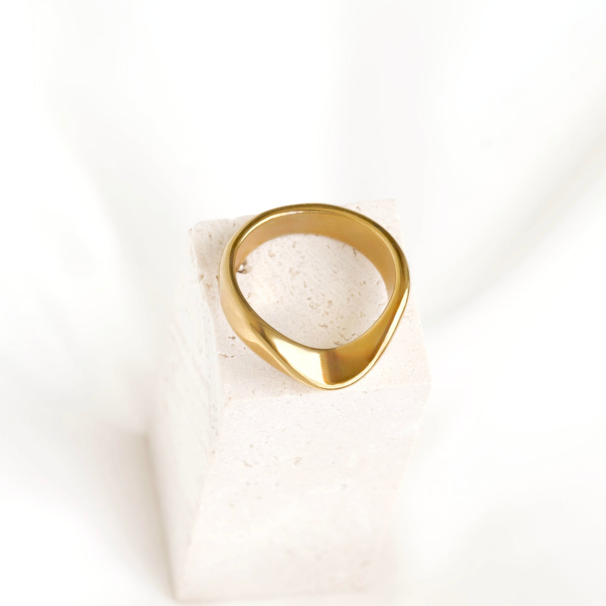 A polished gold signet ring with a wavy, organic shape, displayed on a textured white stone block against a soft white background.
