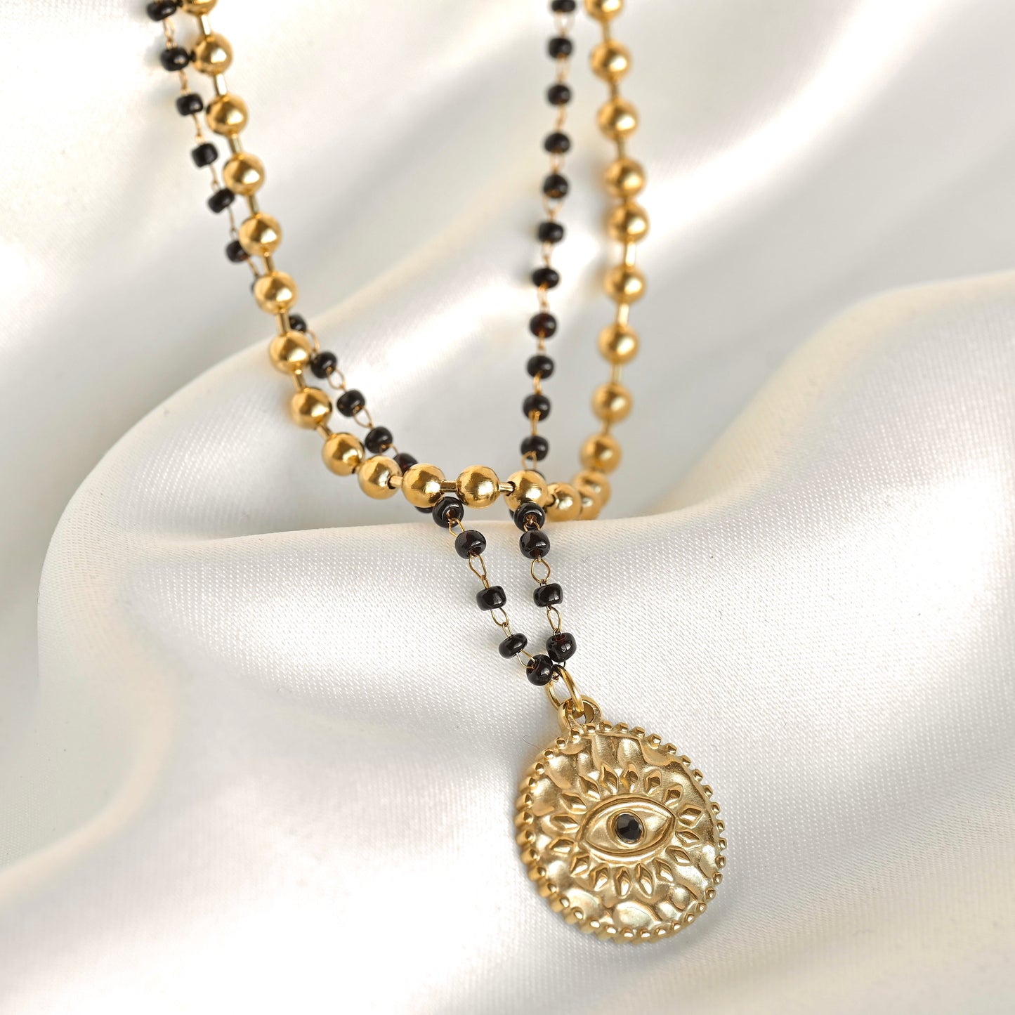 A gold and black beaded necklace with a circular filigree pendant featuring an eye motif, displayed on draped white satin fabric.