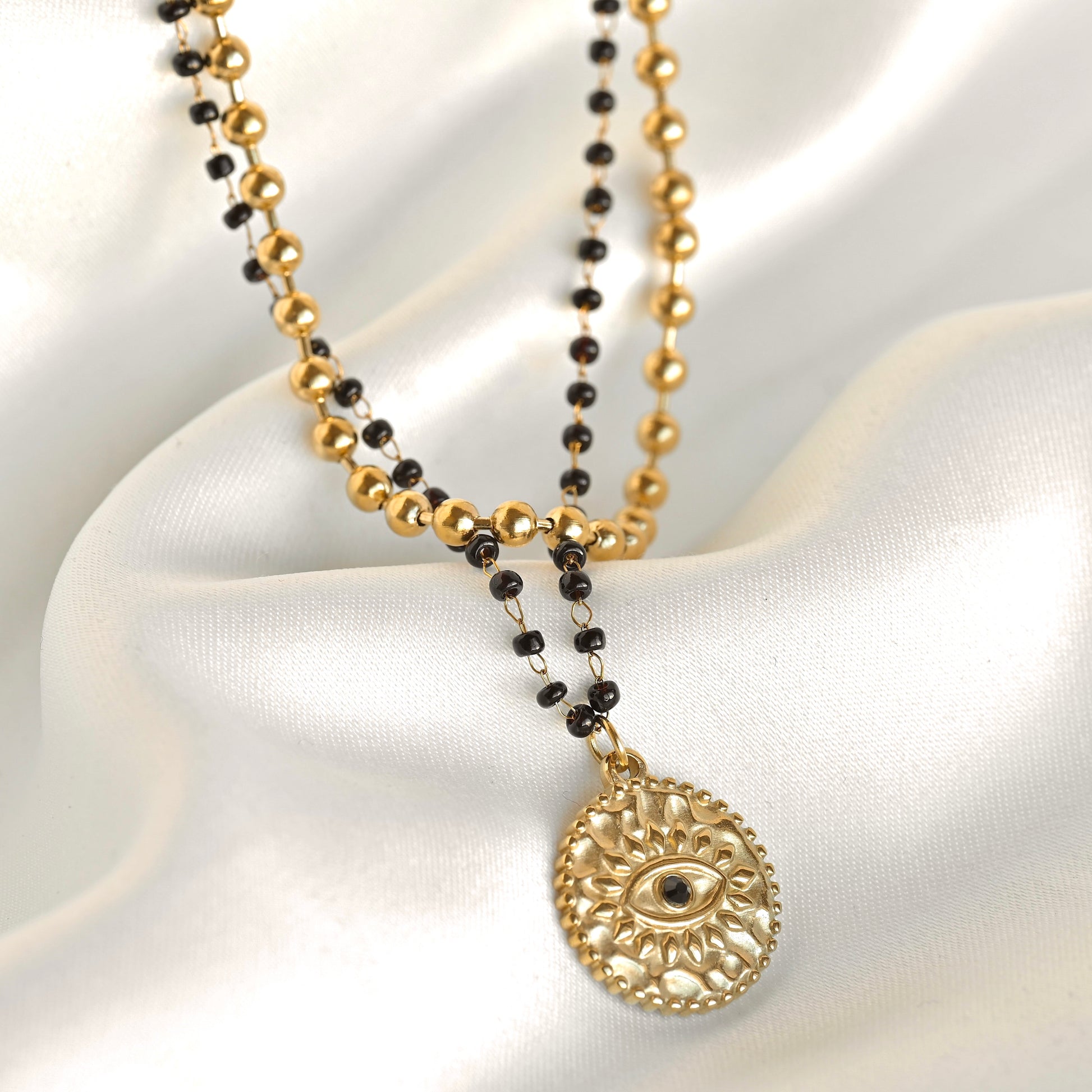 A gold and black beaded necklace with a circular filigree pendant featuring an eye motif, displayed on draped white satin fabric.