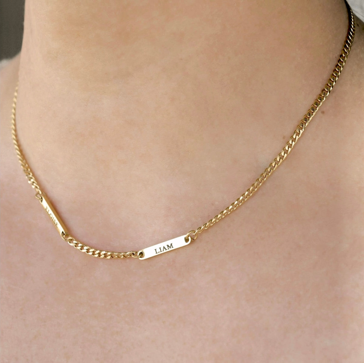 A close-up of a delicate gold curb chain necklace worn on a person's neck, featuring two small rectangular bars engraved with the name 'ELOISE' and 'LIAM'.