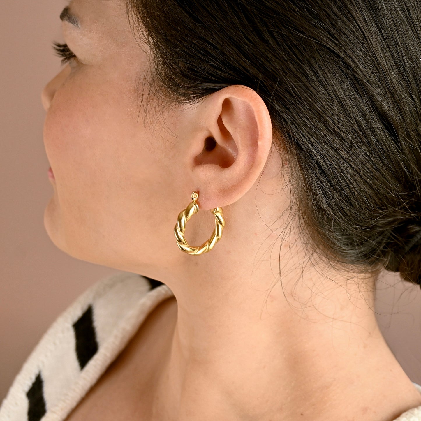 A close-up side profile of a woman wearing a twisted gold hoop earring, showing the earring in detail against a soft beige background.