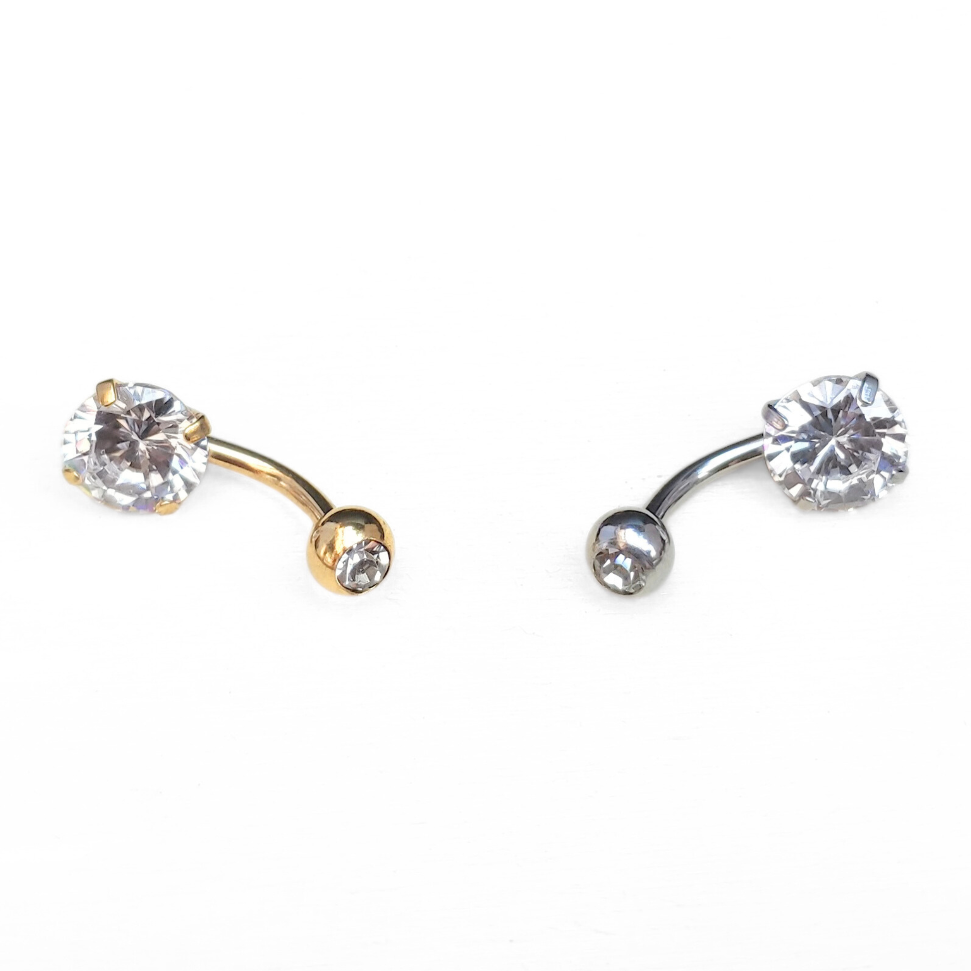 Two belly button rings, one gold one silver, with clear gemstones in a 4 prong setting on a white background