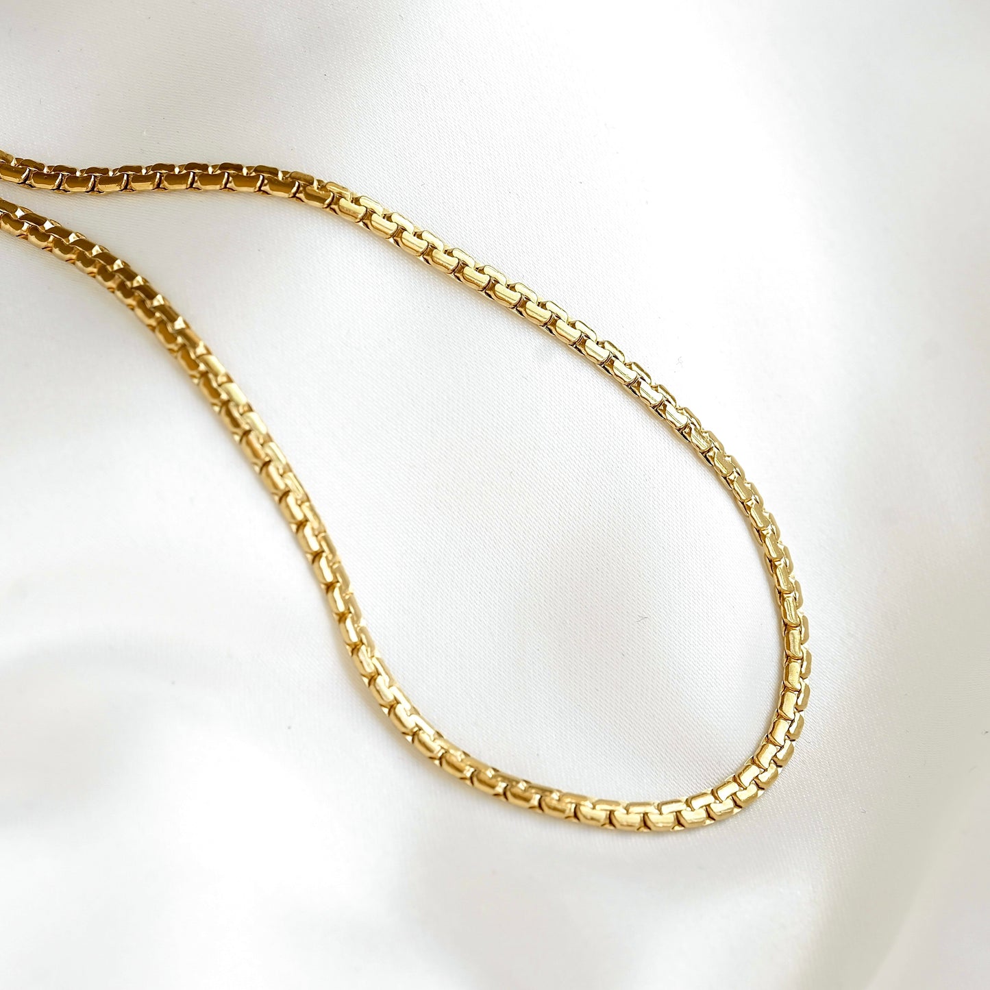 A gold-colored curb chain necklace draped in an oval shape on a white satin fabric background.