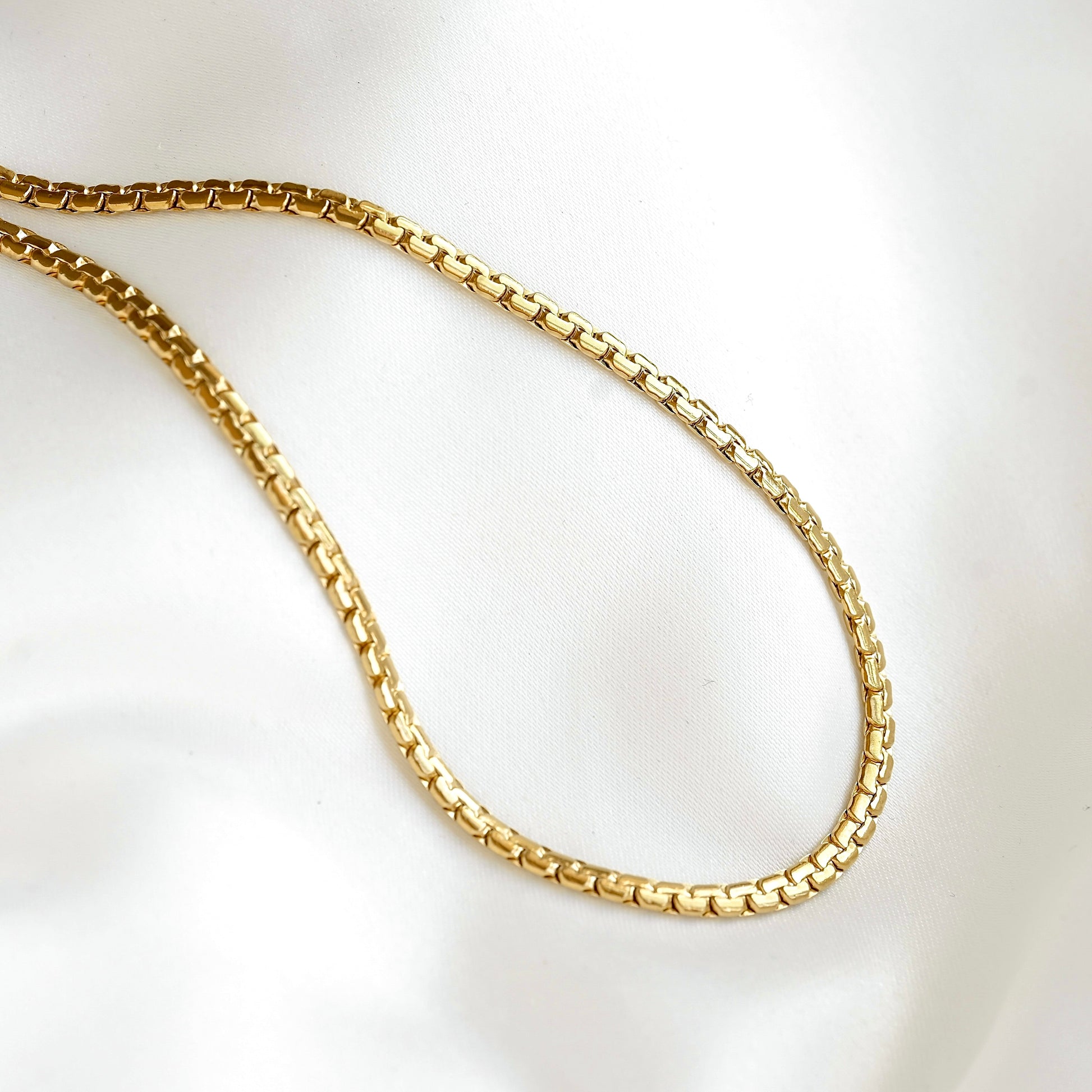 A gold-colored curb chain necklace draped in an oval shape on a white satin fabric background.