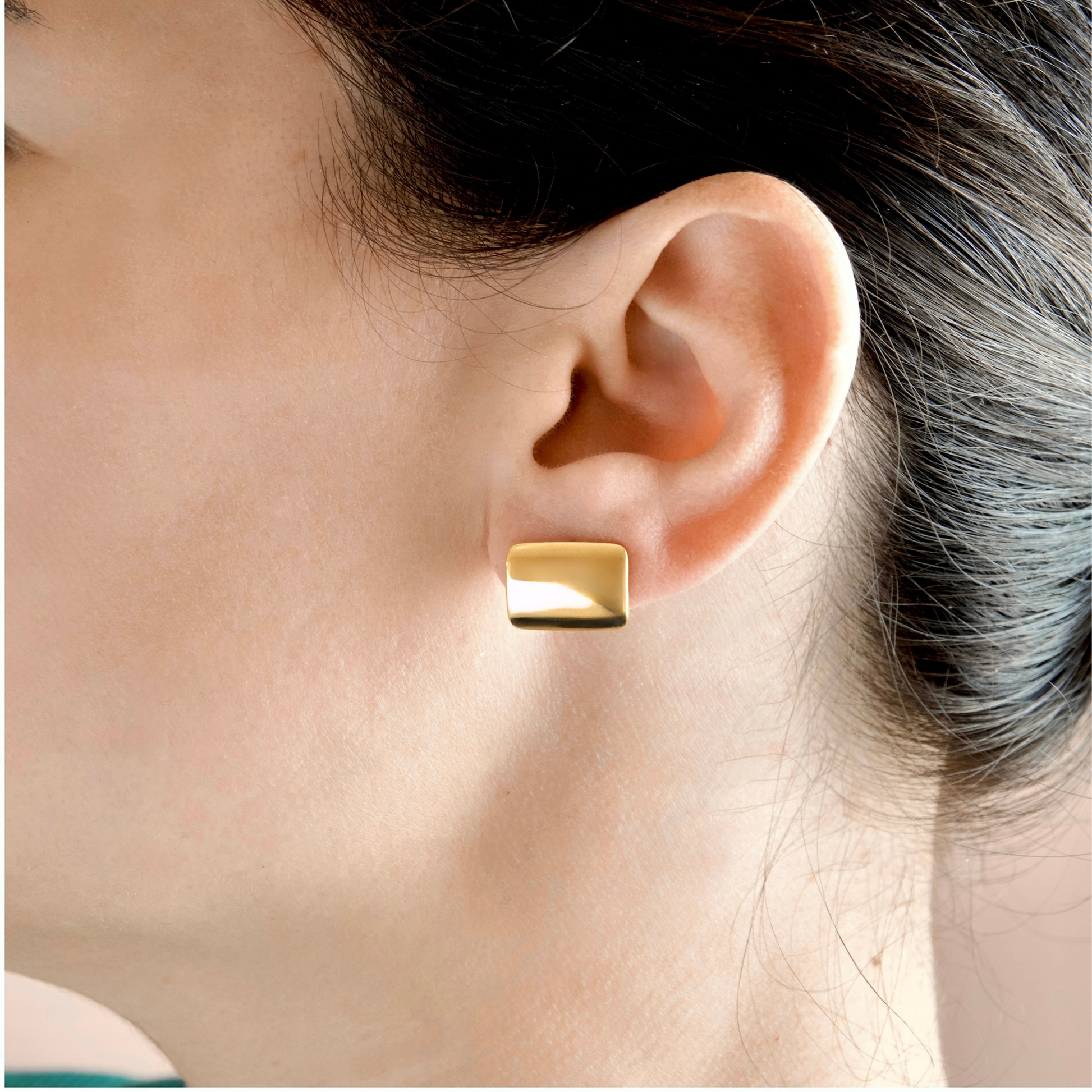 A close-up of a person's ear wearing a small rectangular gold stud earring with a polished finish.