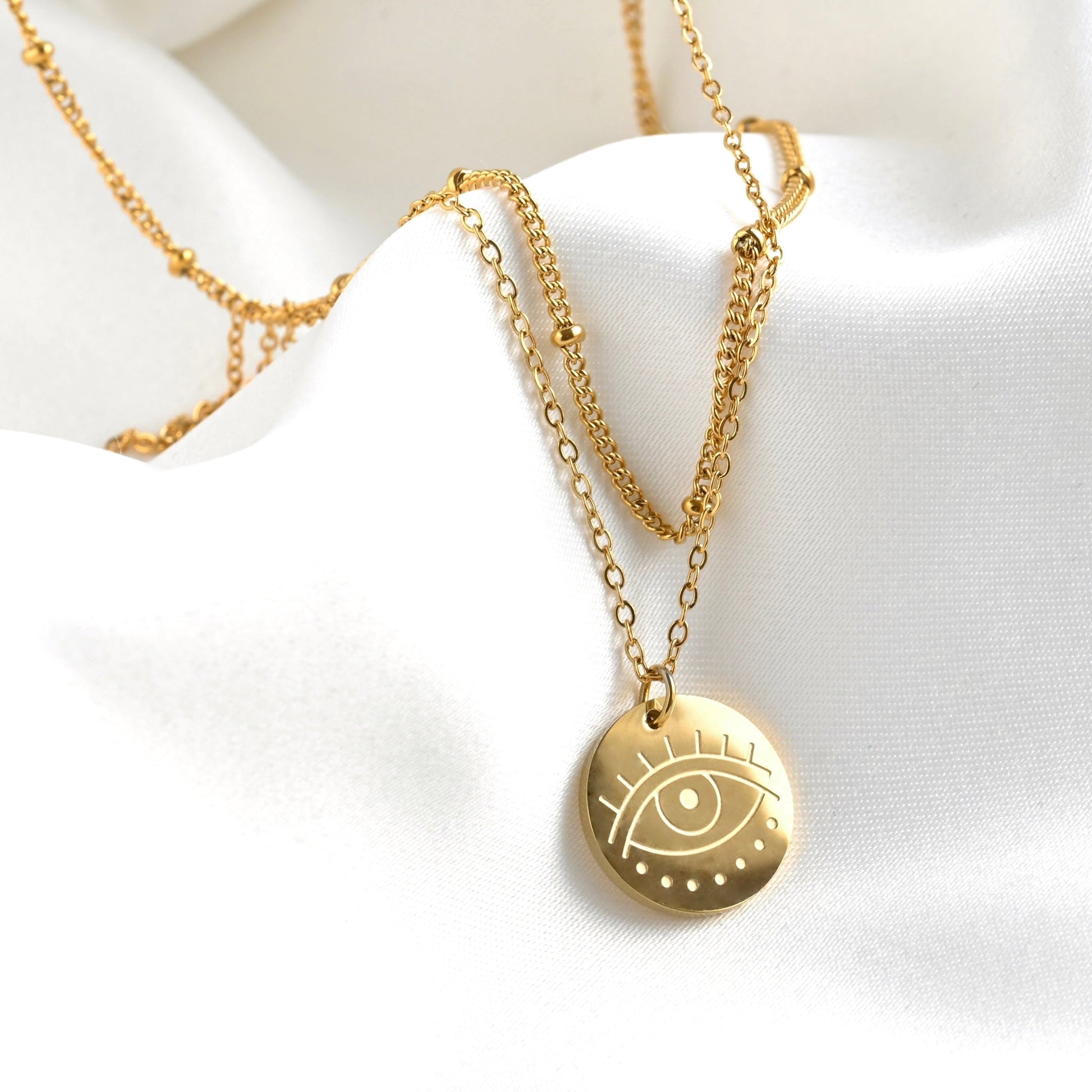 Front view of layered gold necklace with an eye coin pendant on a white fabric background