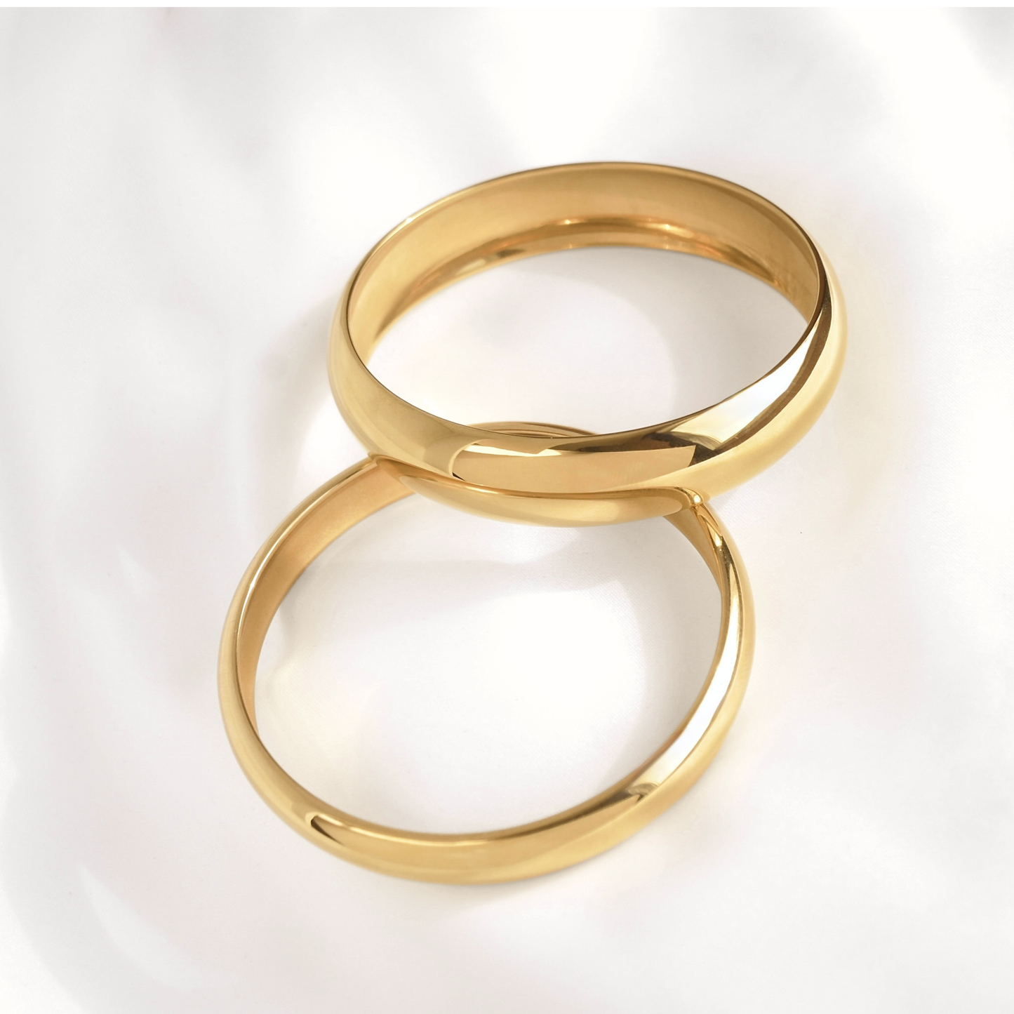 Two polished gold bangle bracelets with a smooth, rounded design, overlapping on a soft white background.