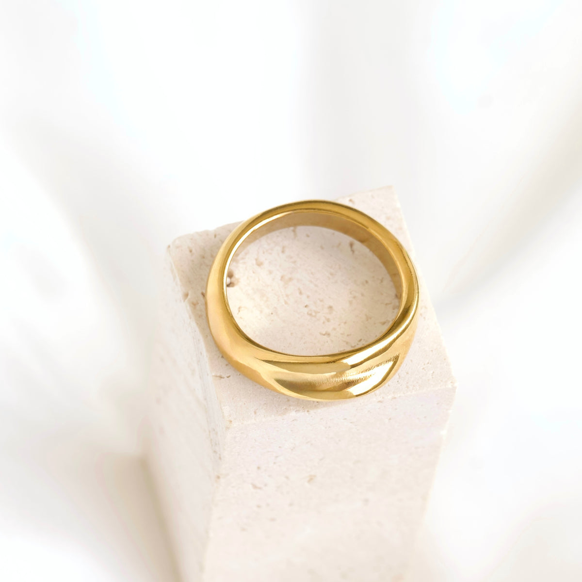 A polished gold ring with a wide, sculptural band displayed on a textured beige stone block against a soft white background.