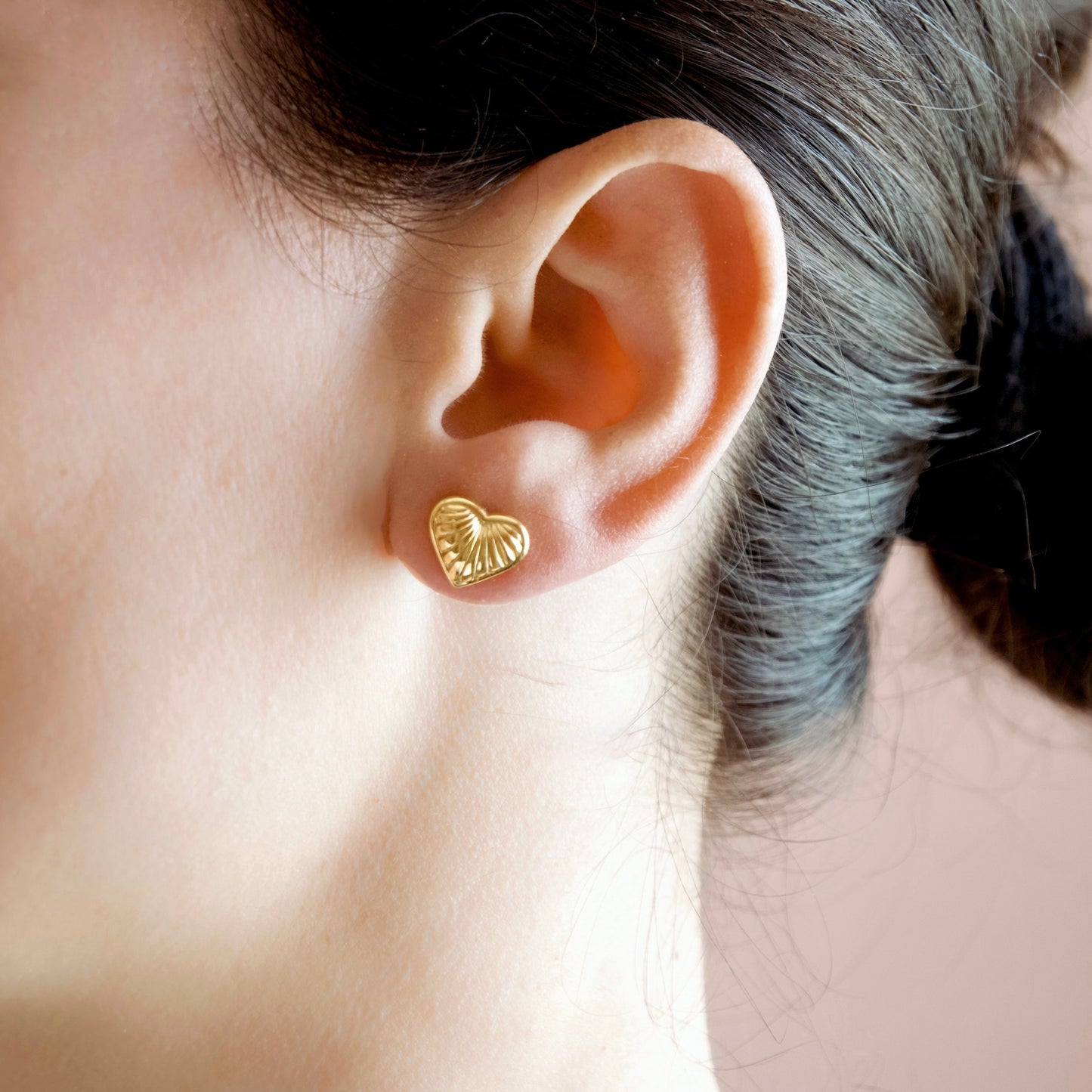 A close-up of a person's ear wearing a small textured gold heart stud earring.