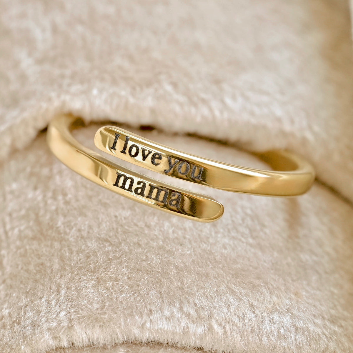 A gold wrap-style ring with engraved text reading 'I love you mama' on a soft beige textured fabric background.