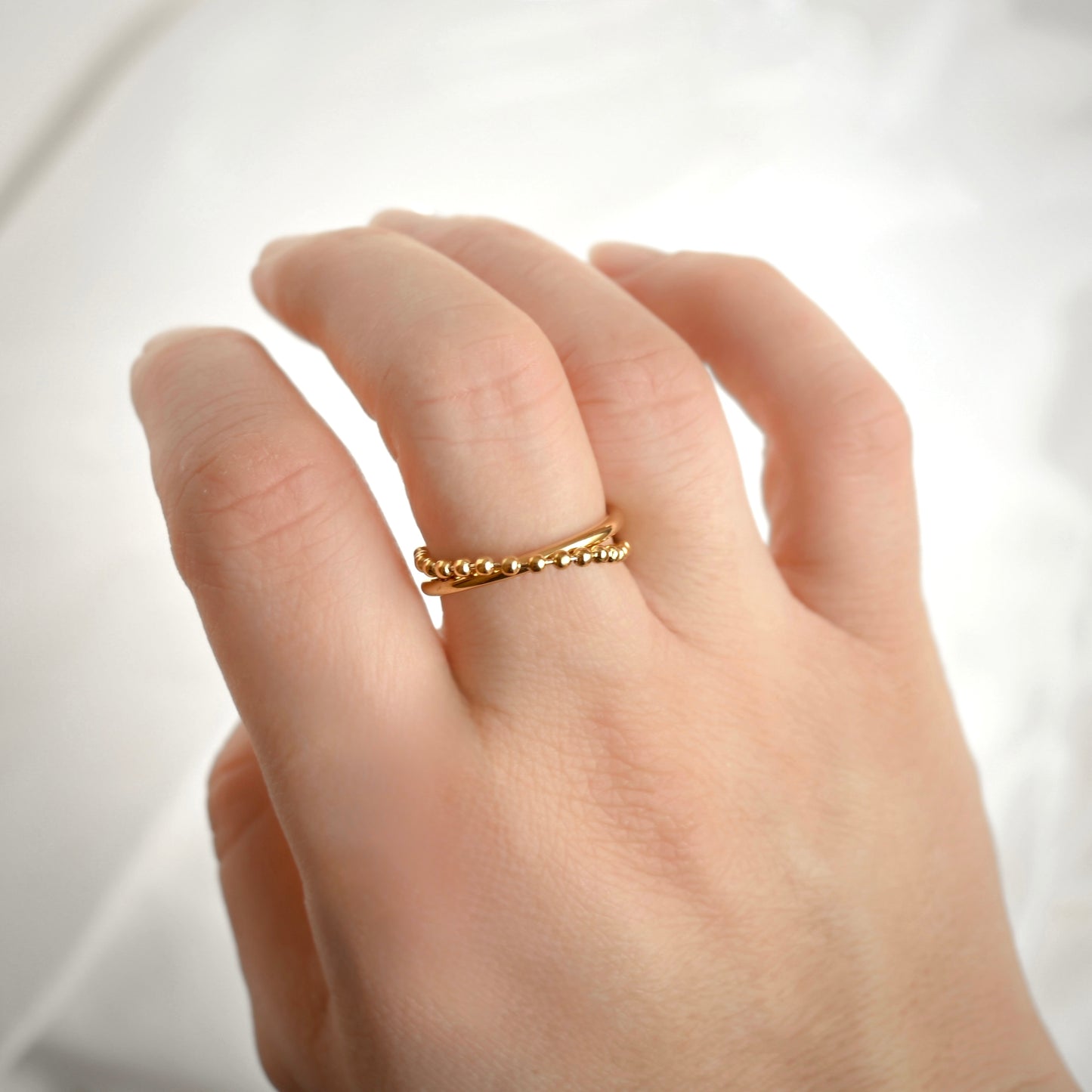 A close-up of a hand wearing a delicate gold X ring with a textured beaded chain design, set against a soft, out-of-focus white background.