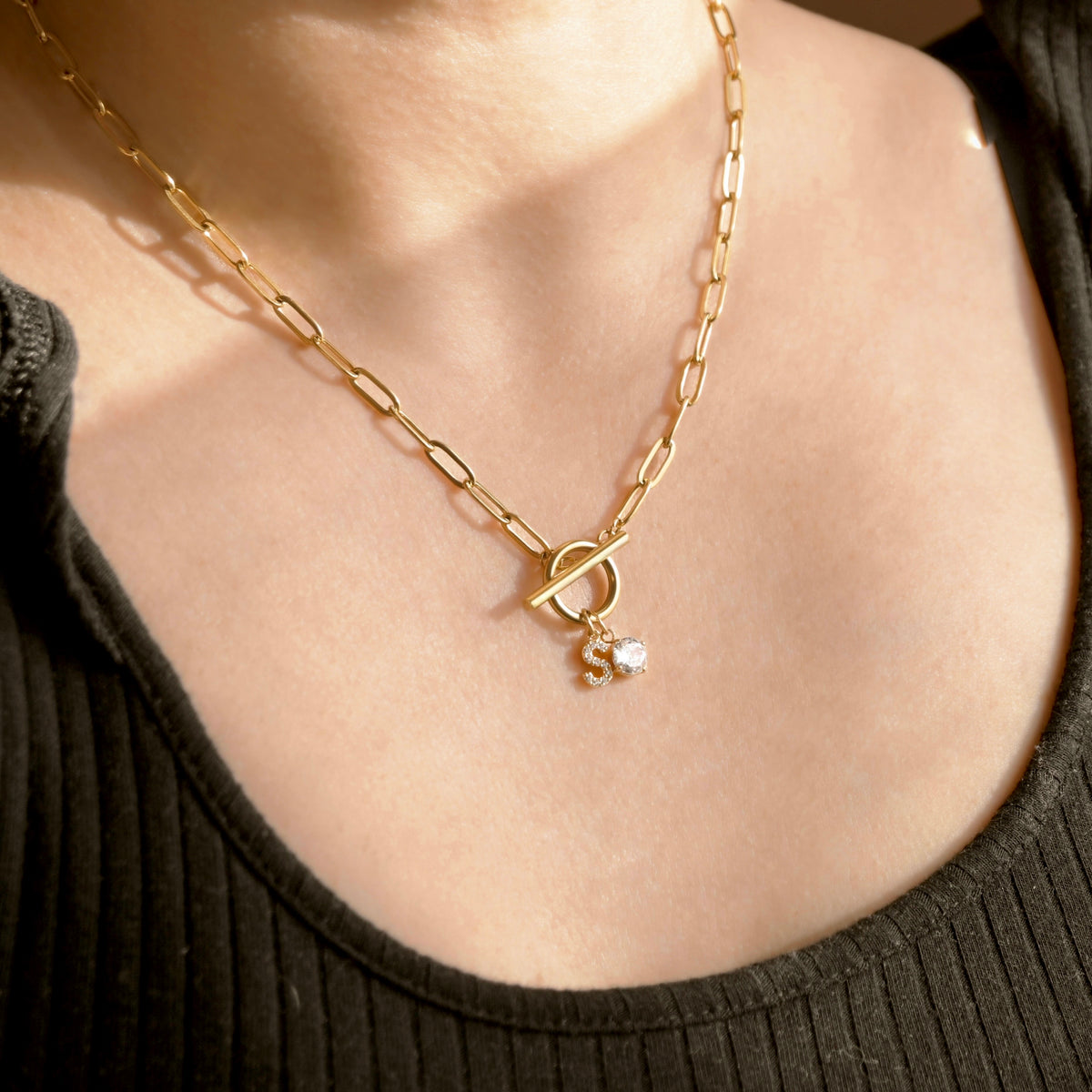 A close-up of a gold  paperclip chain necklace with a toggle clasp and a small dangling charm, worn on a model's neck over a dark ribbed top.