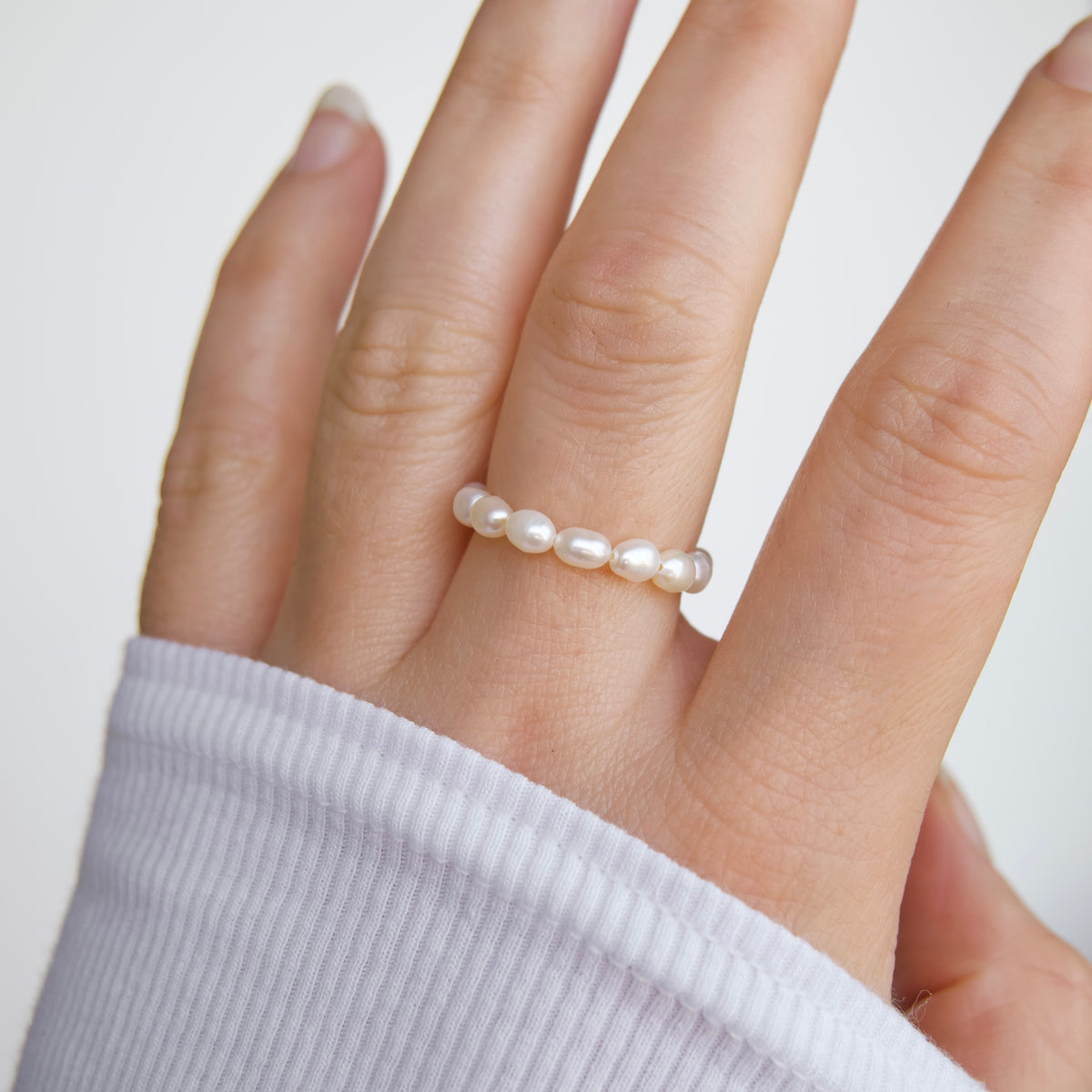 A close-up of a hand wearing a delicate ring made of five small, round, lustrous white pearls spaced evenly around a thin band, styled with a white ribbed knit sleeve.
