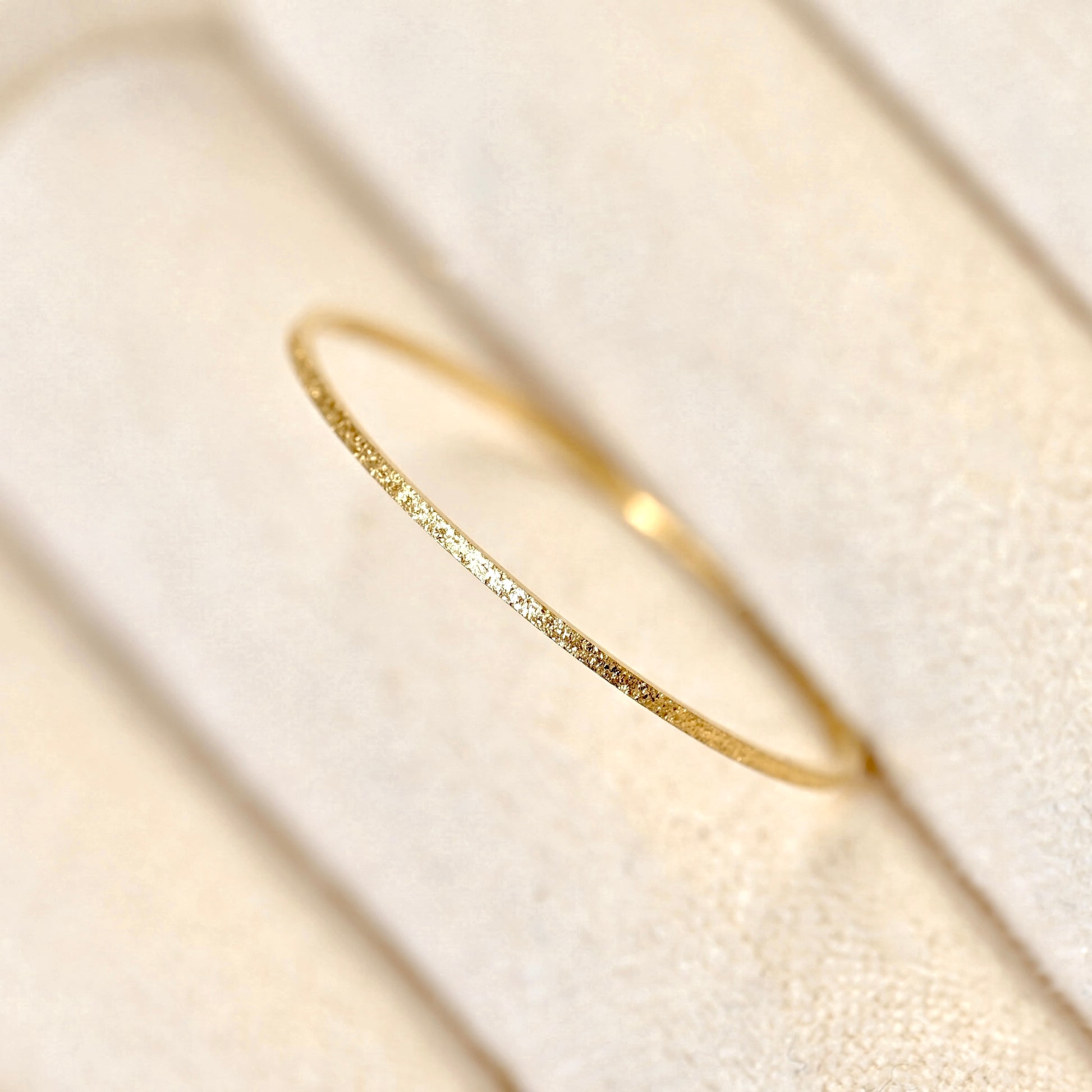 A delicate, thin gold ring with a textured, hammered finish, displayed on a soft beige textured ring stand.