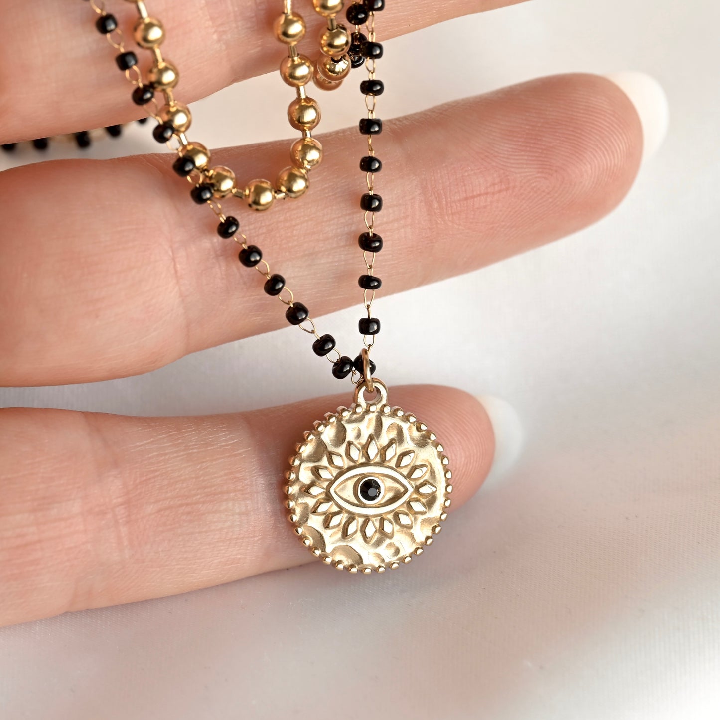 A gold-toned circular pendant necklace with an eye motif hangs from a beaded chain worn on a person's hand. The chain alternates between polished gold beads and small black beads, shown against a light neutral background.