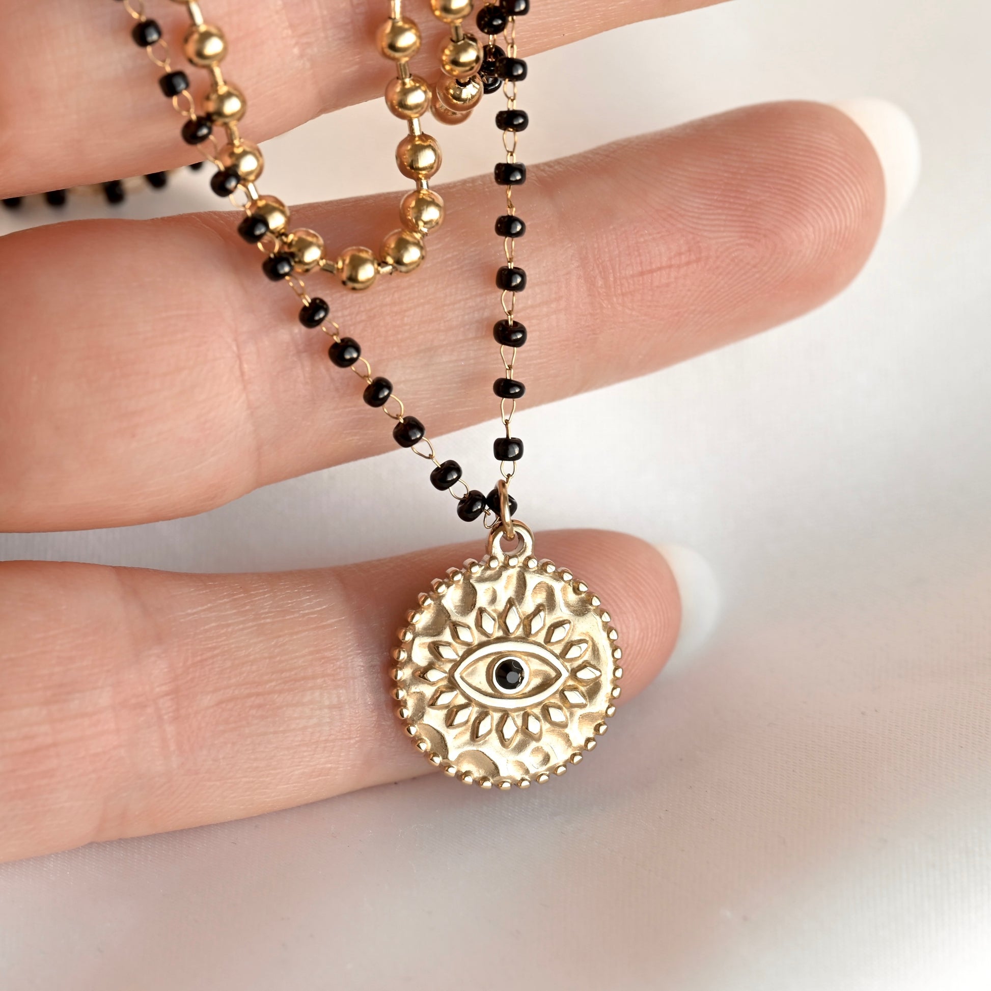 A gold-toned circular pendant necklace with an eye motif hangs from a beaded chain worn on a person's hand. The chain alternates between polished gold beads and small black beads, shown against a light neutral background.