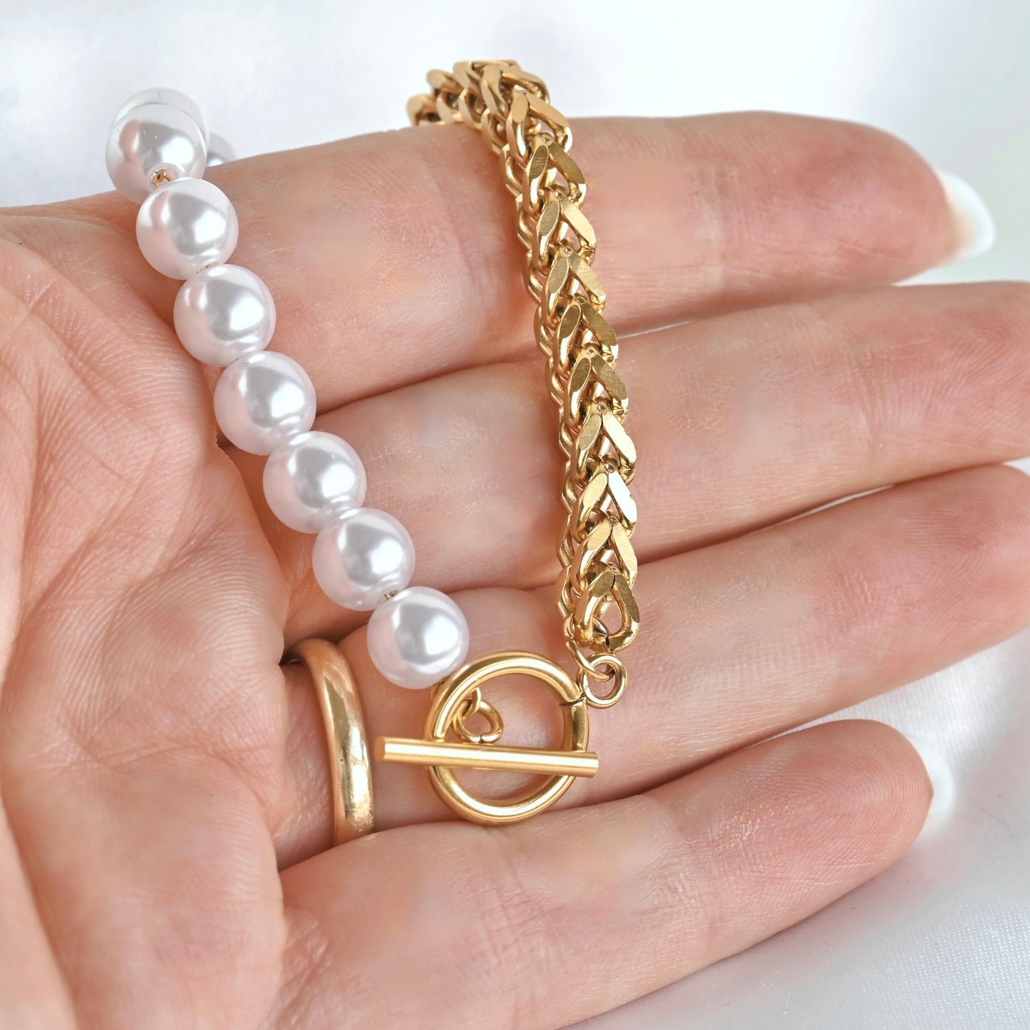 A close-up of a hand displaying a gold chain bracelet with a toggle clasp paired with a strand of white pearl beads.
