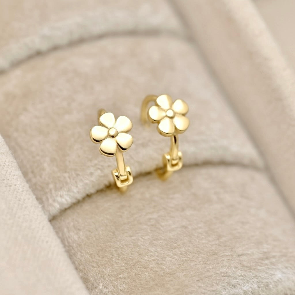 A pair of small gold hoop earrings with flower-shaped tops, displayed on a beige velvet jewelry box.