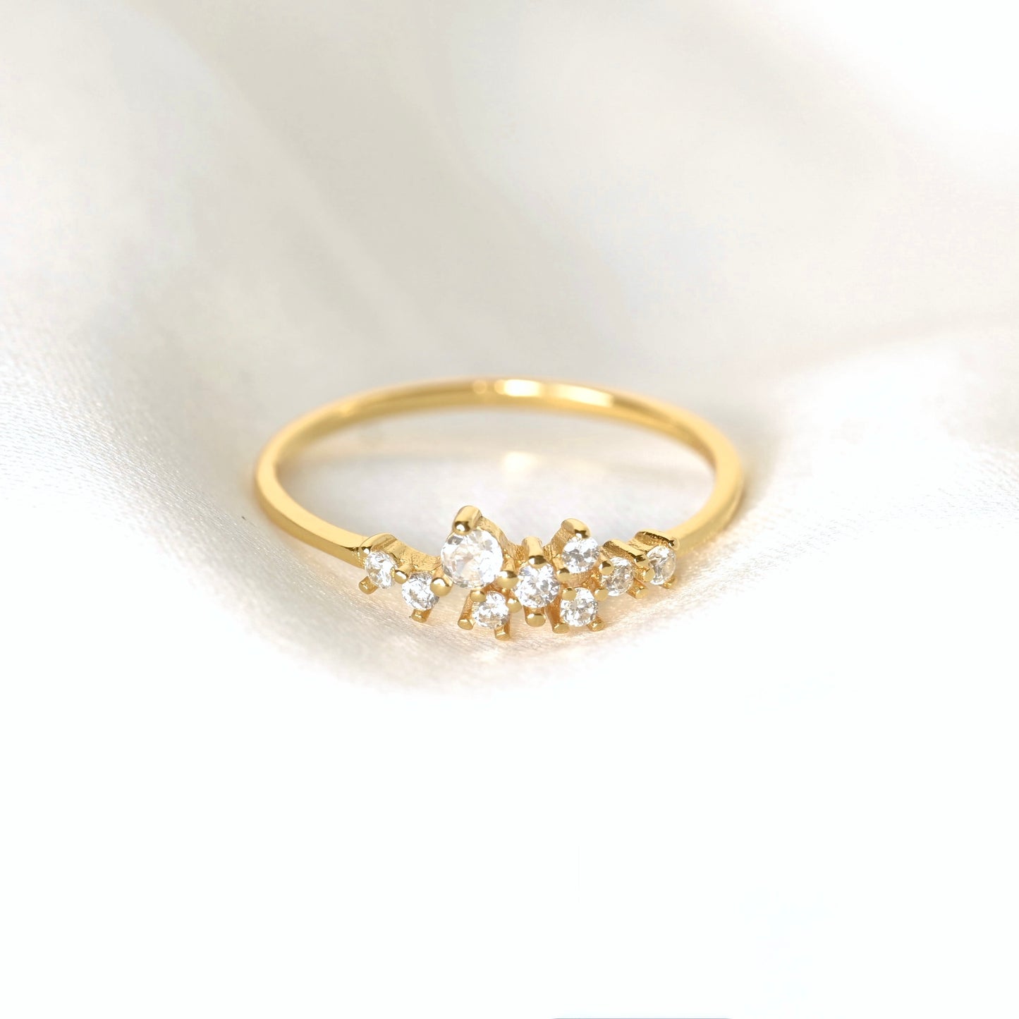 A delicate gold ring featuring a small cluster of clear round-cut CZ stones in a floral or leaf-like arrangement, resting on a soft white fabric background.