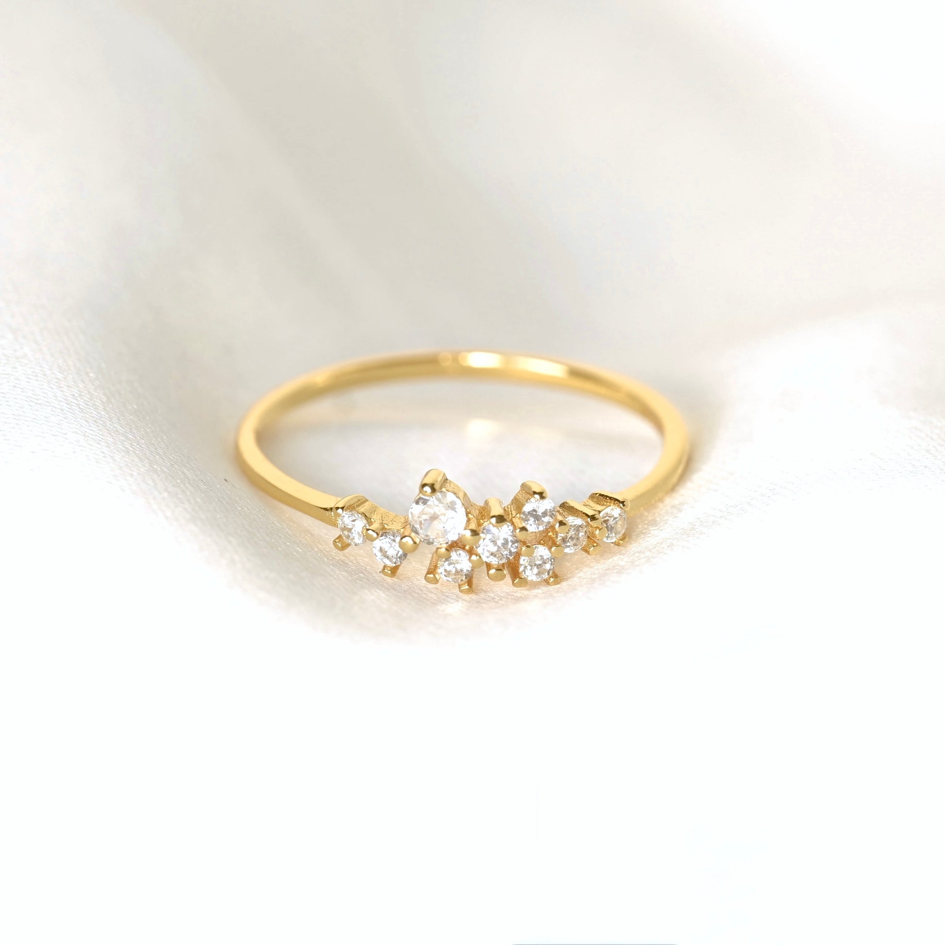 A delicate gold ring featuring a small cluster of clear round-cut CZ stones in a floral or leaf-like arrangement, resting on a soft white fabric background.