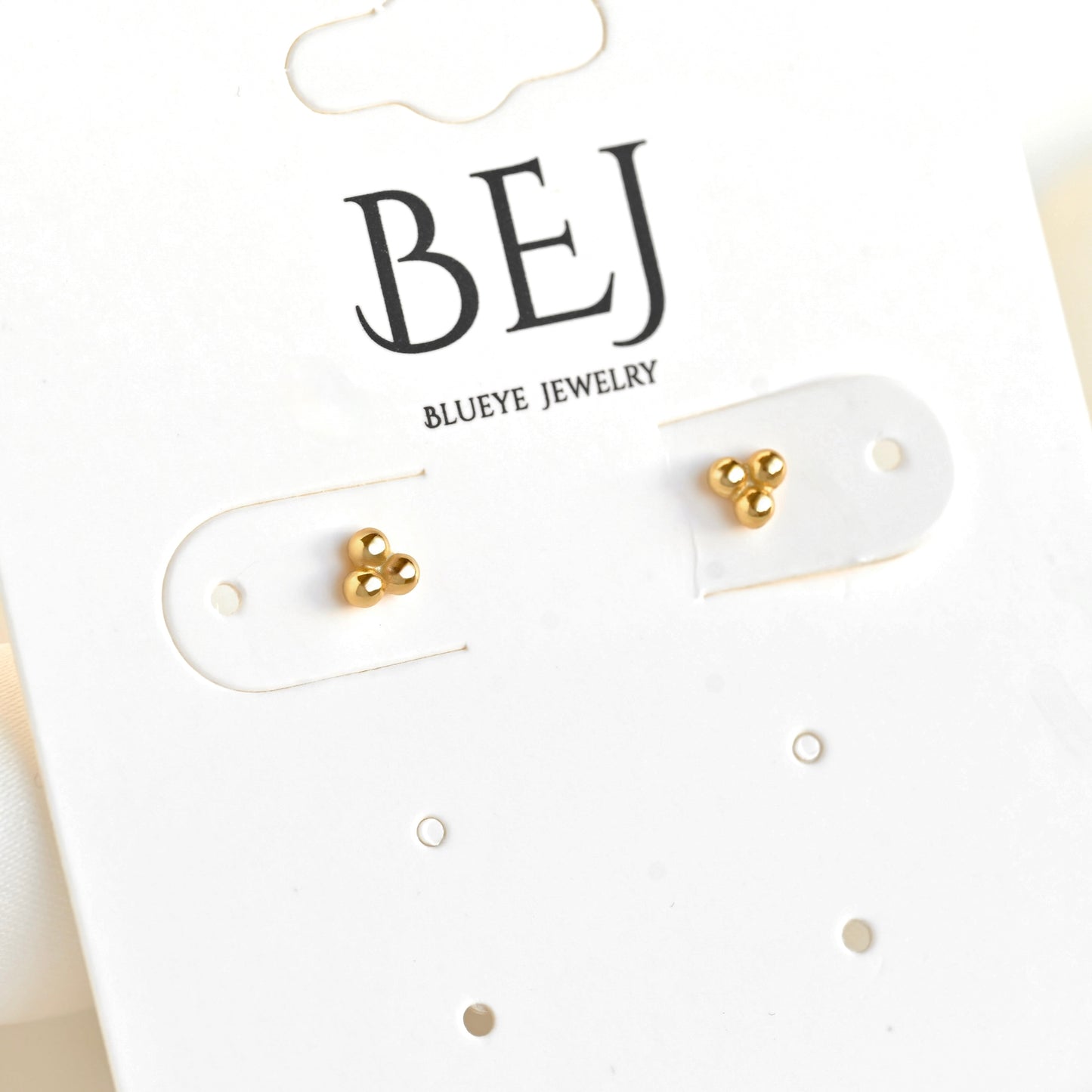A pair of small gold stud earrings displayed on a white BEJ Blueeye Jewelry card.