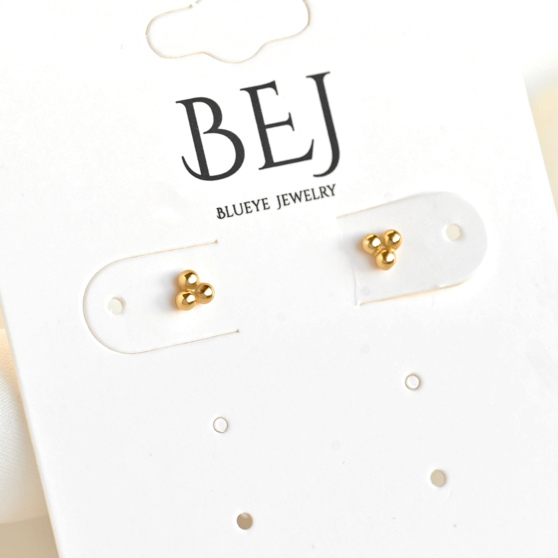 A pair of small gold stud earrings displayed on a white BEJ Blueeye Jewelry card.