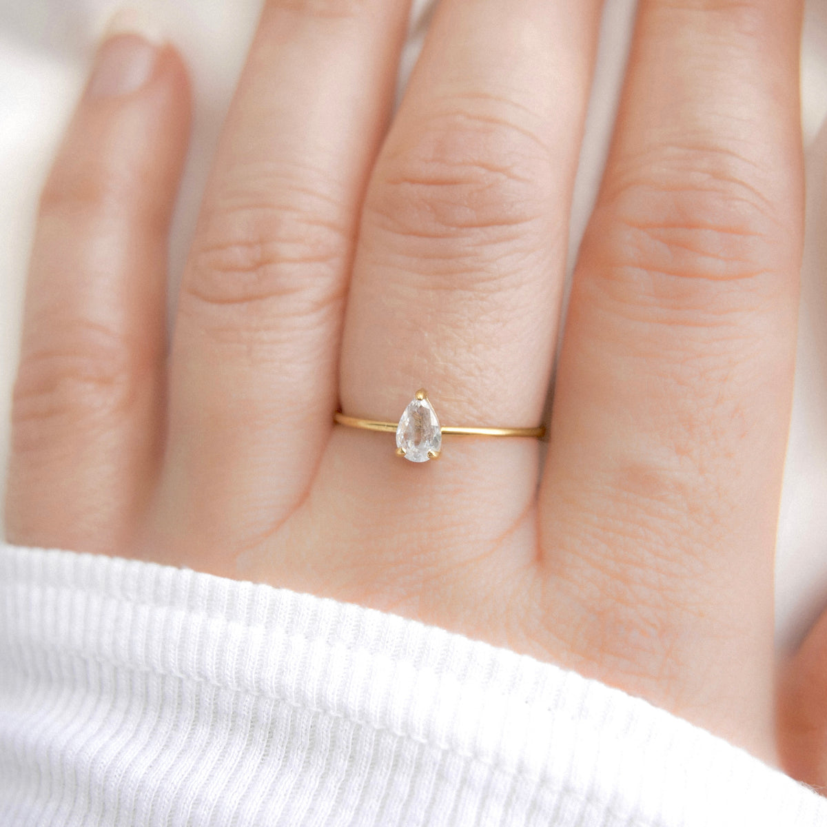 A close-up of a hand wearing a delicate gold ring with a pear-cut clear gemstone, set against a soft white background.