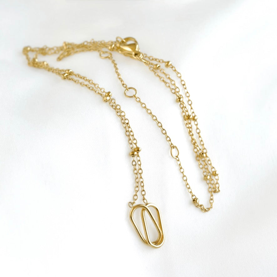 A delicate gold chain necklace with a small abstract open-loop pendant, displayed on a soft white fabric background.