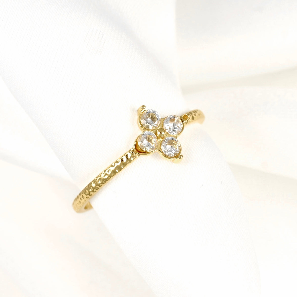 A gold ring featuring a small four-stone floral cluster with clear round-cut gemstones, displayed on a soft white fabric ring holder.