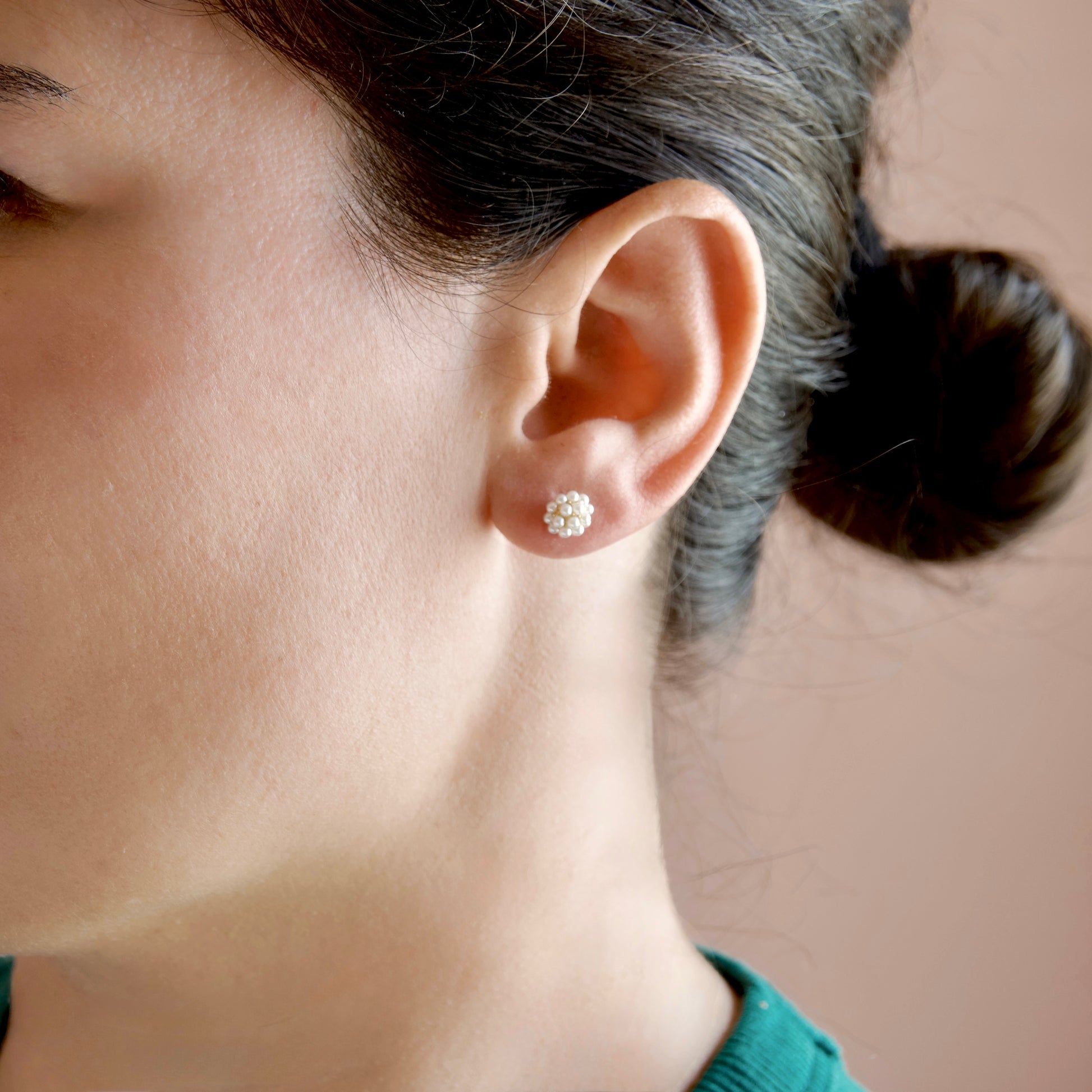 A close-up of a person's ear wearing a small pearl cluster ball stud earring made of milky white round pearls in a gold-tone setting.