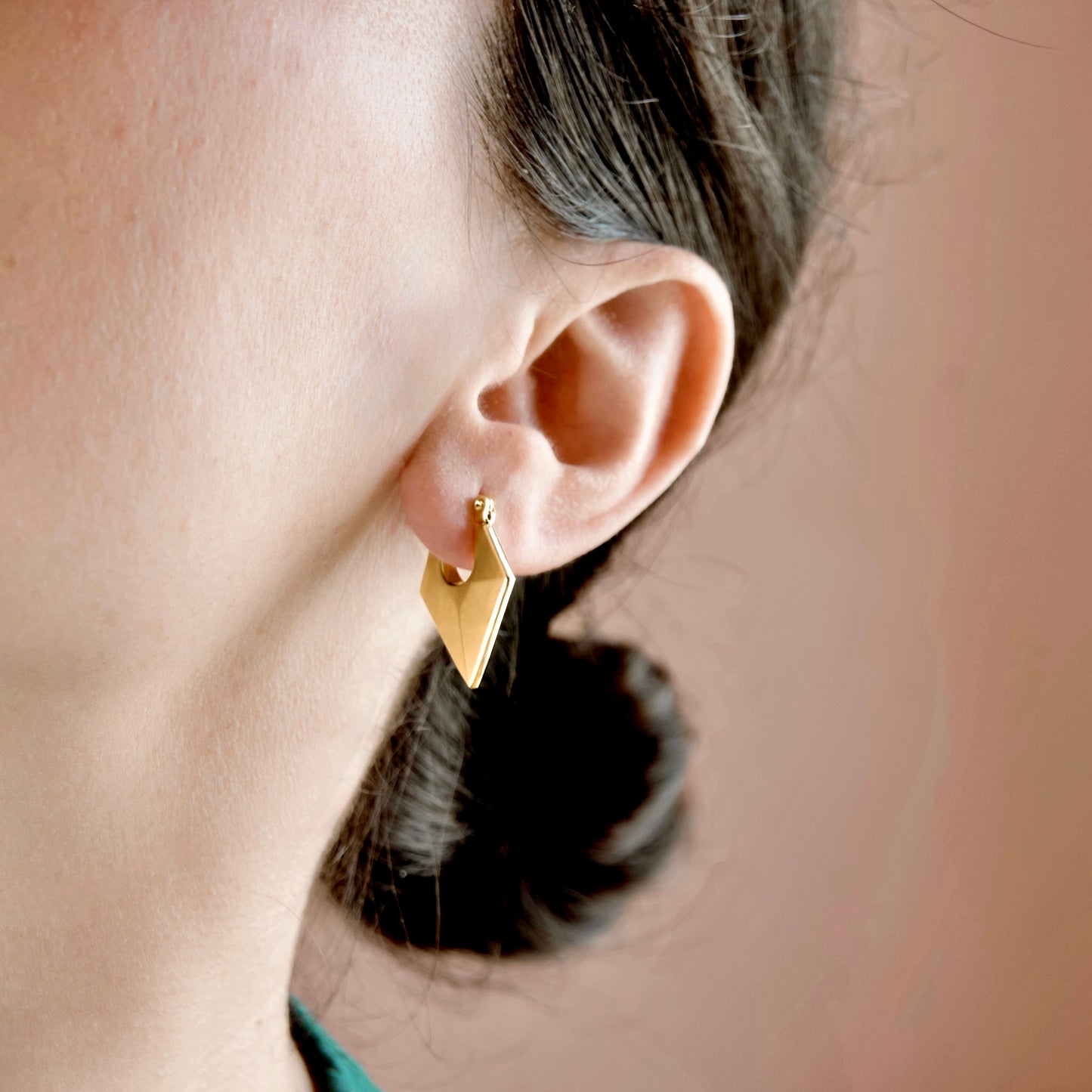 A close-up of a person's ear wearing a small gold geometric dangle earring against a soft peach background.