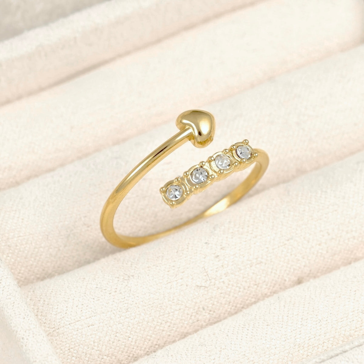 A gold open-band heart ring with a small curved bar set with four clear round stones, displayed on a cream-colored textured jewelry cushion.