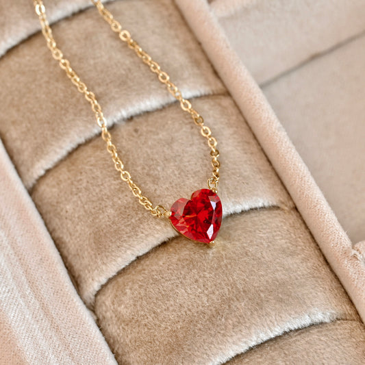 Gold necklace with a red heart-shaped pendant on a beige textured surface
