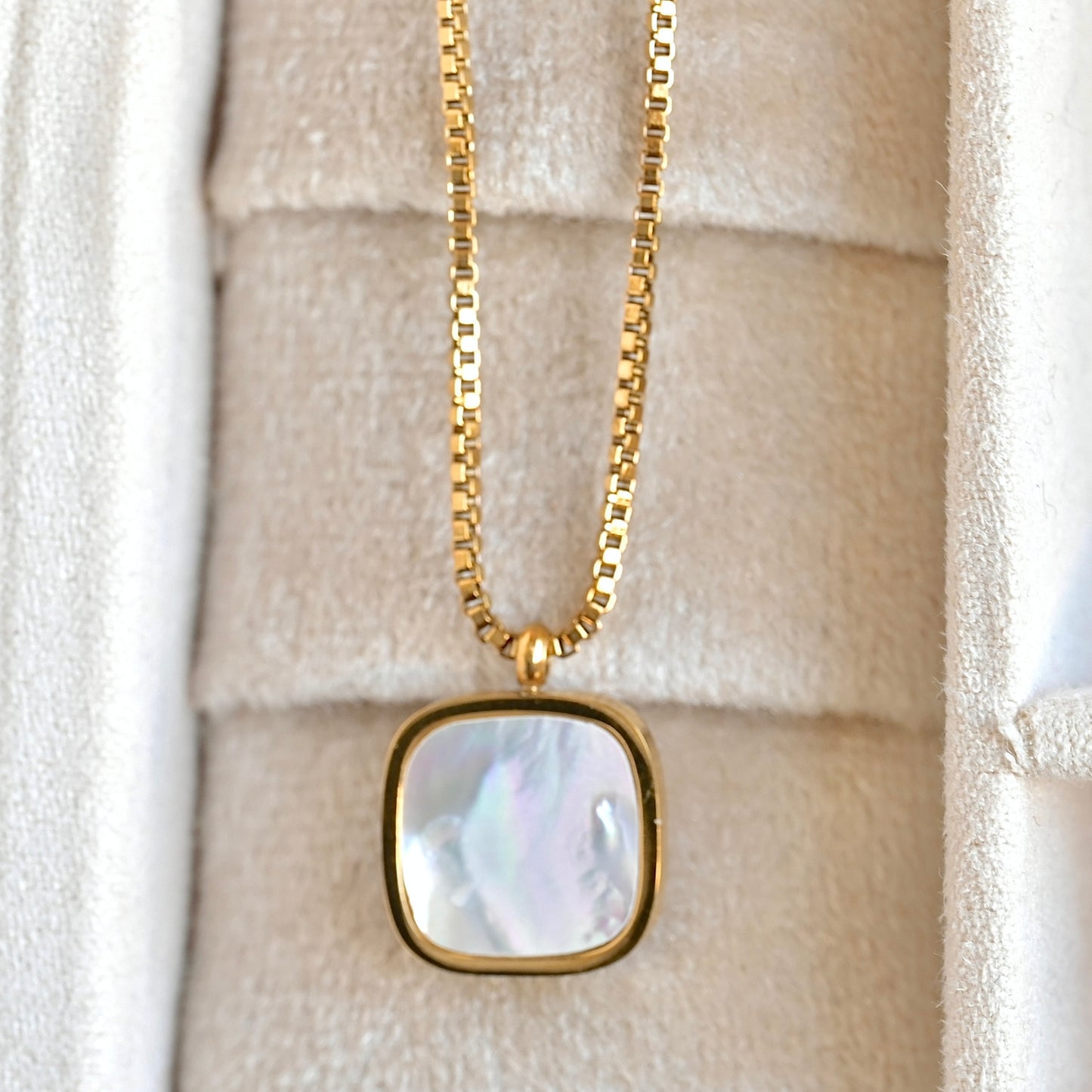 A gold box chain necklace with a square pendant featuring a pearlescent white and iridescent shell inlay, displayed inside a cream-colored jewelry box.