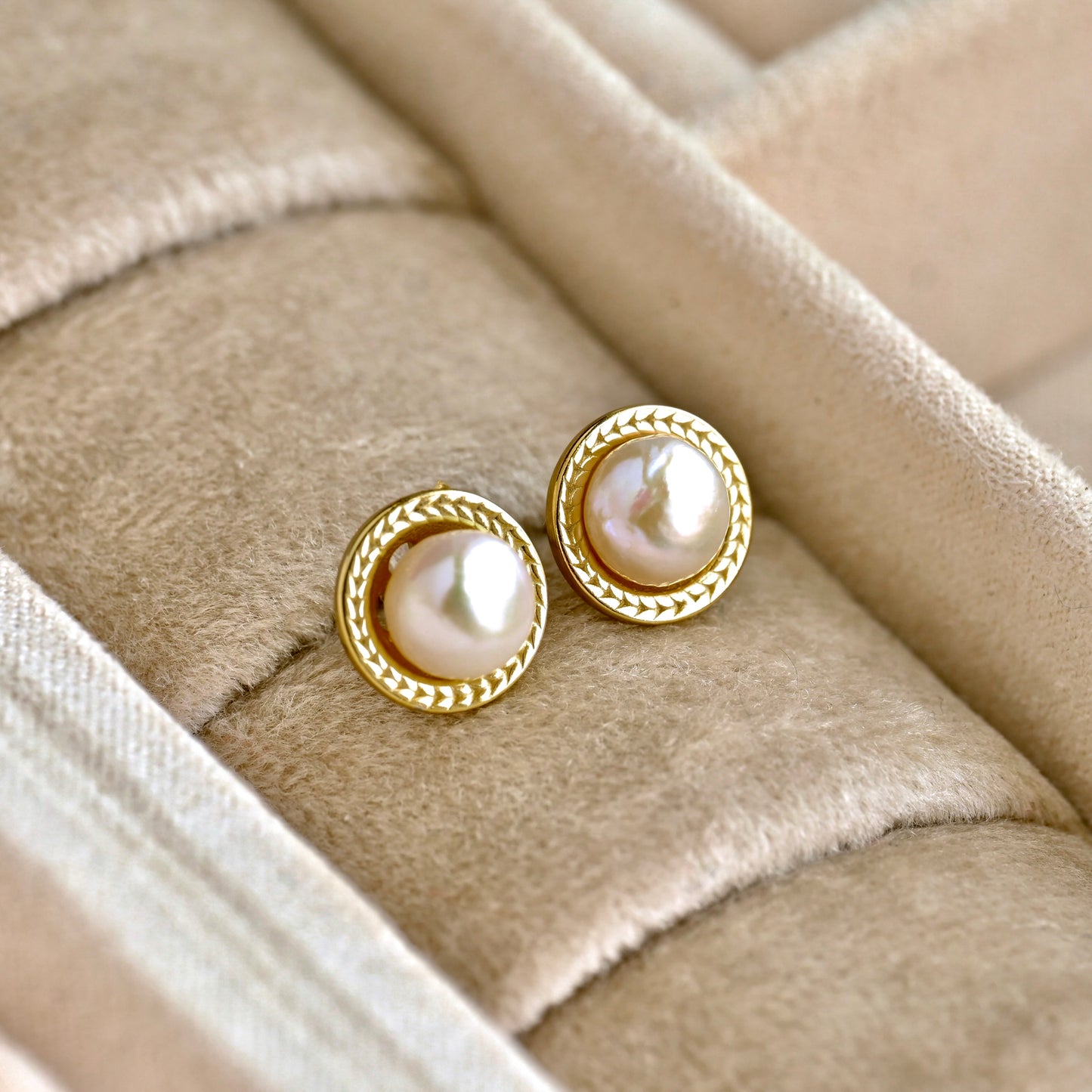 A pair of gold-toned stud earrings with round white pearls and a braided rope border, displayed on a beige velvet jewelry box.