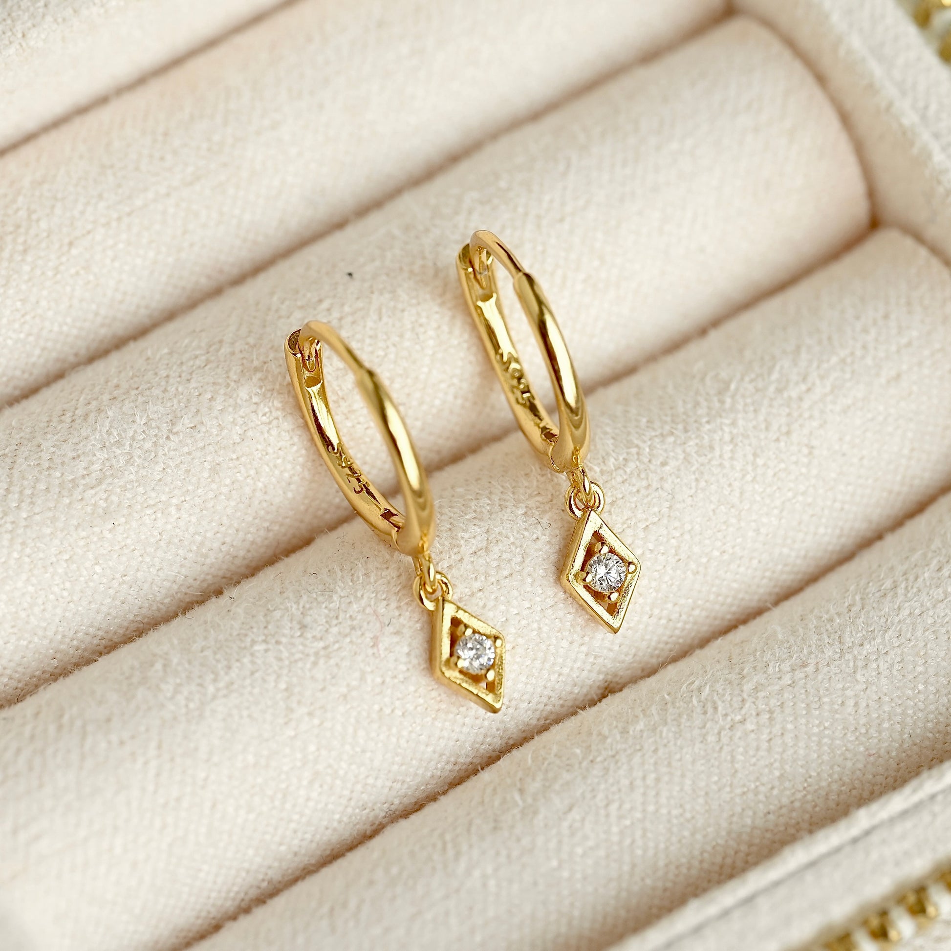 A pair of gold huggie hoop earrings with dangling diamond-shaped charms set with clear stones, displayed on a cream-colored jewelry box cushion.