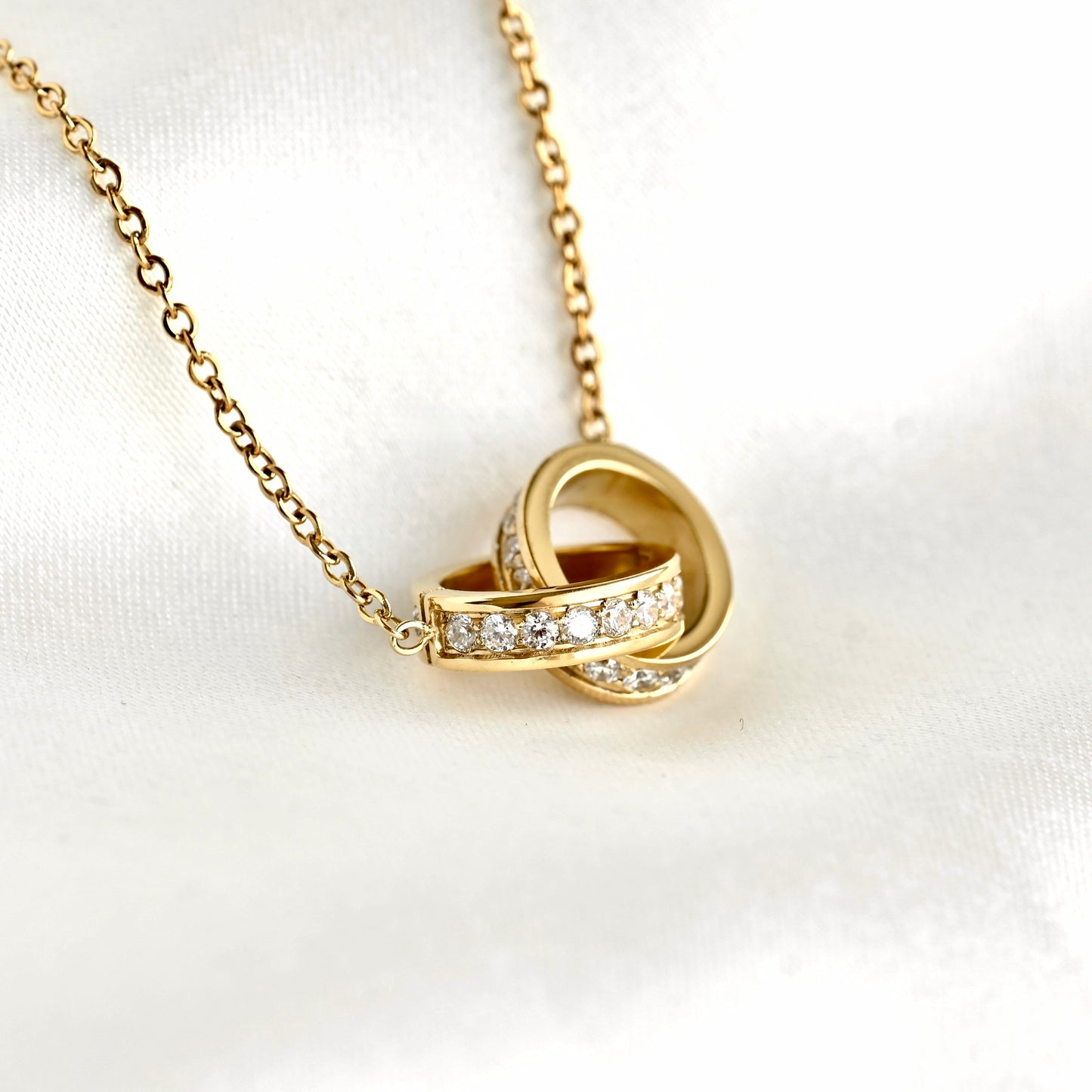A gold chain necklace with an interlocking knot pendant, featuring a row of clear pavé stones across the band,s displayed on a white satin fabric background.