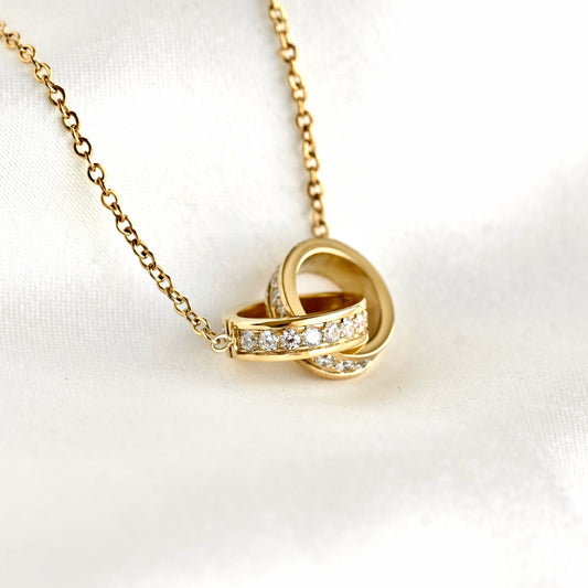 A gold chain necklace with an interlocking knot pendant, featuring a row of clear pavé stones across the band,s displayed on a white satin fabric background.