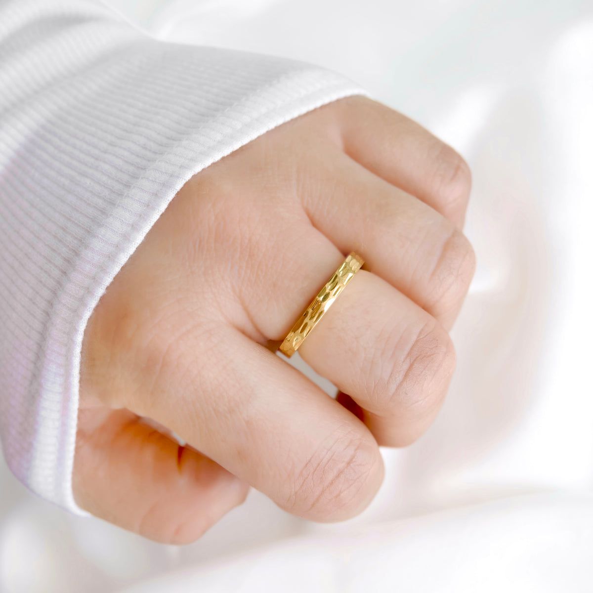 A close-up of a hand wearing a textured yellow gold band ring, shown against a soft white background with a white ribbed sleeve visible.