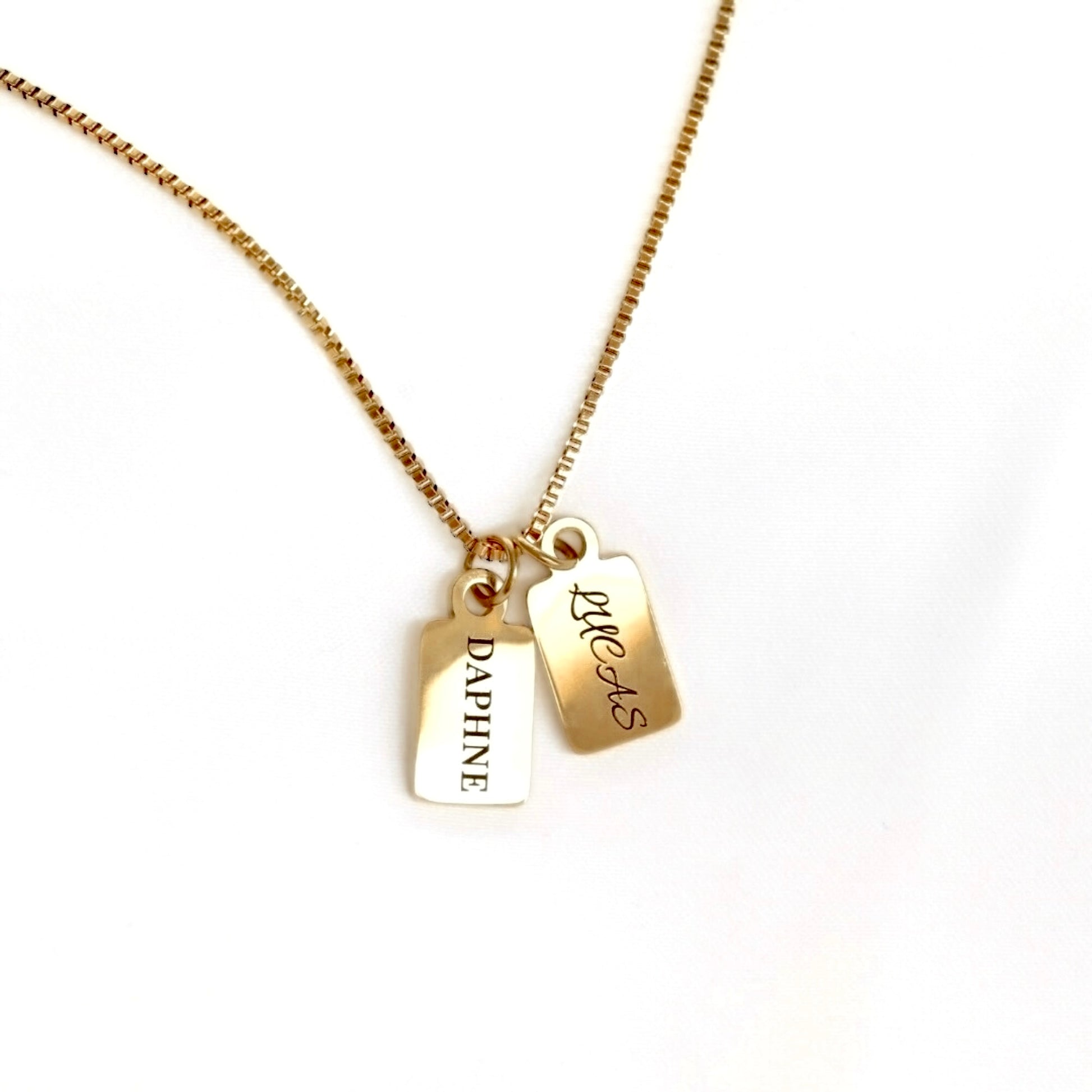 A gold chain necklace with two small rectangular pendant tags hanging from a gold box chain. One tag is engraved with the name 'DAPHNE', while the other is  engraved with cursive 'LUCAS'.