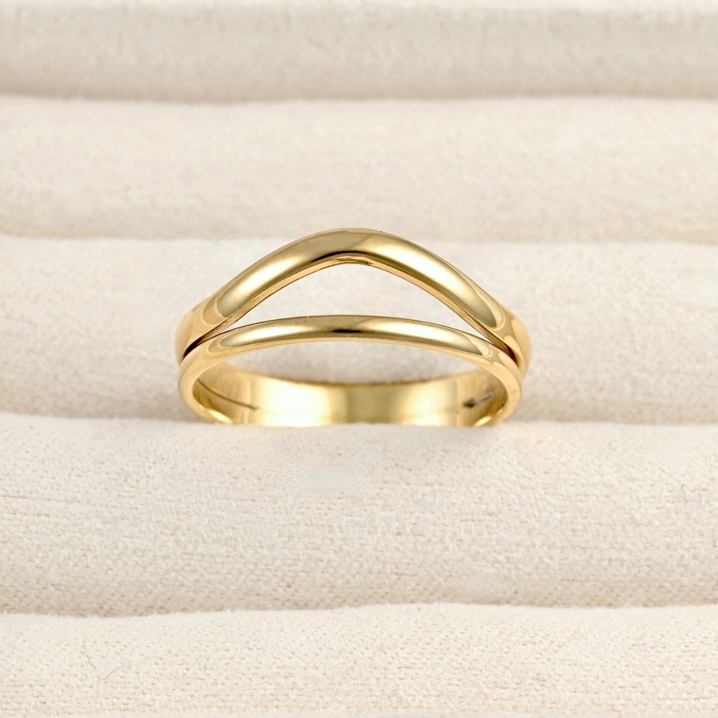 A polished gold ring with a double curved, interlocking design, displayed on a textured cream-colored fabric surface.