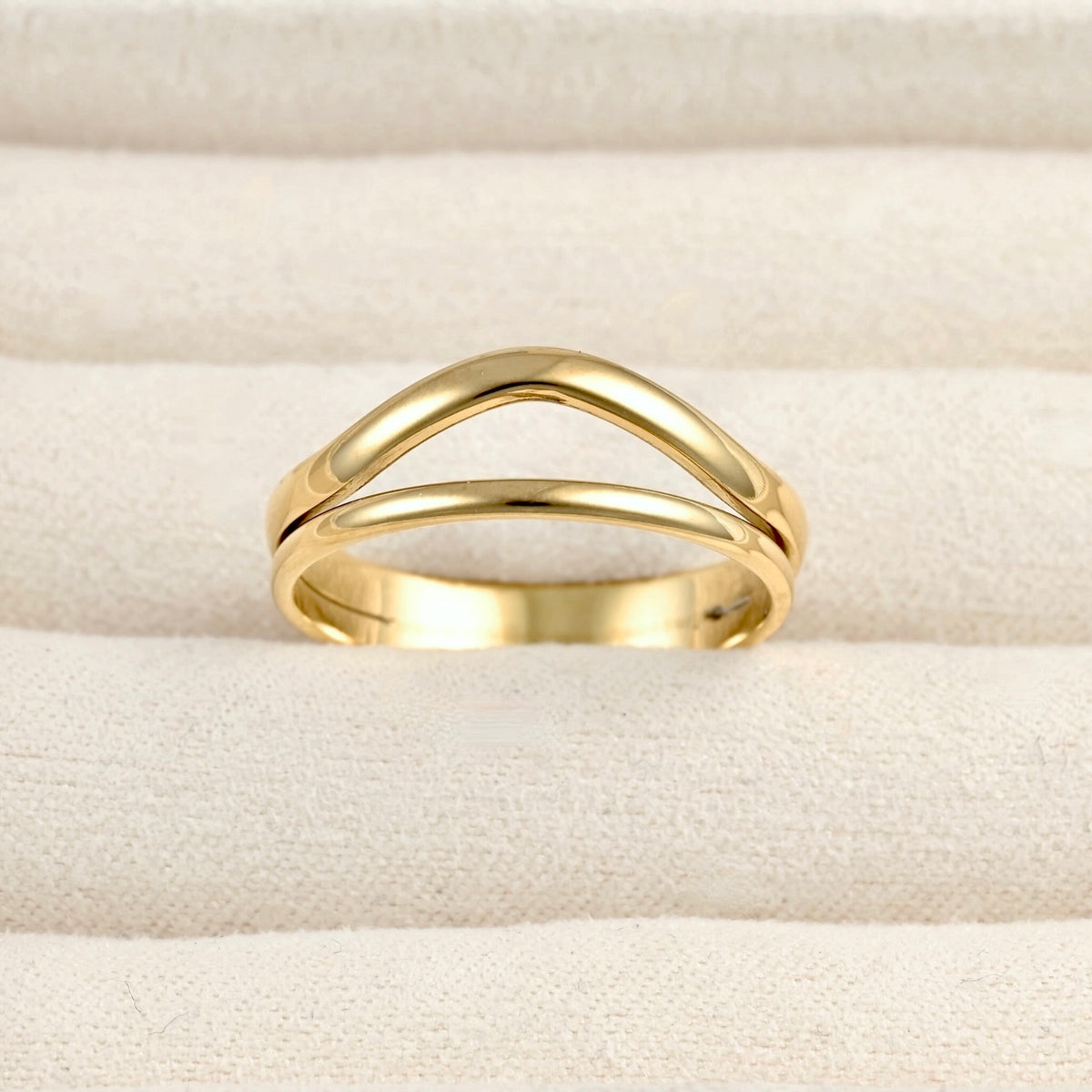 A polished gold ring with a double curved, interlocking design, displayed on a textured cream-colored fabric surface.