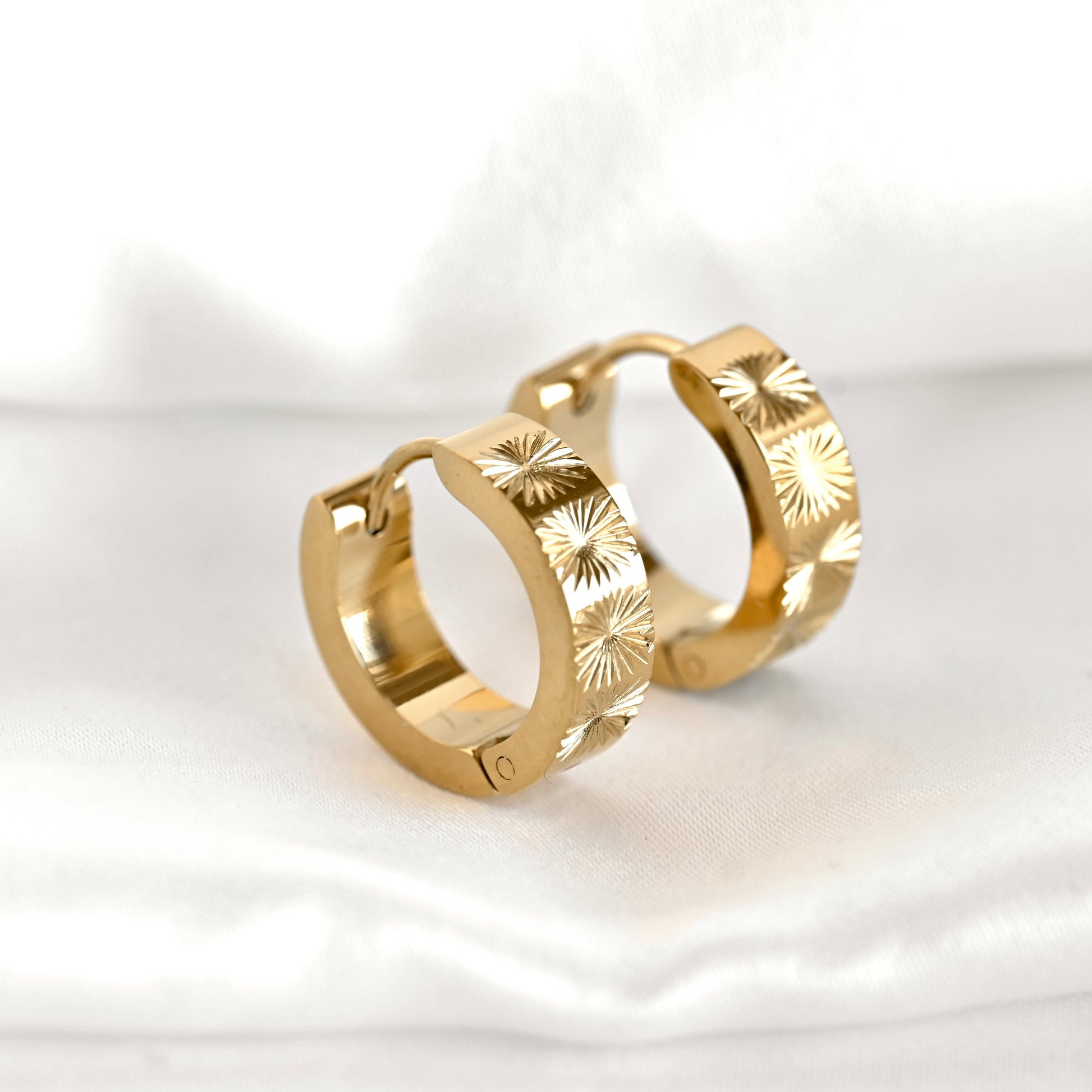 A pair of gold huggie hoop earrings with a sunburst engraved pattern on the outer band, resting on a white satin fabric background.