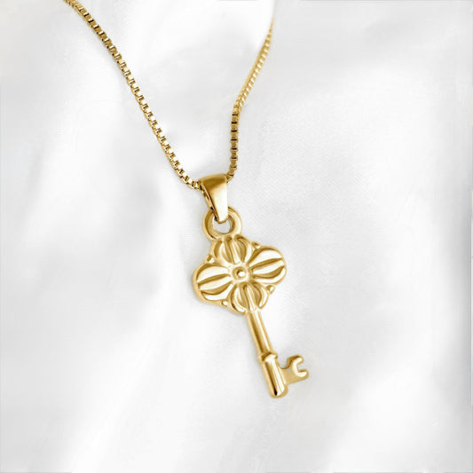 A gold key-shaped pendant necklace with a flower-like top, displayed on a white satin fabric background.