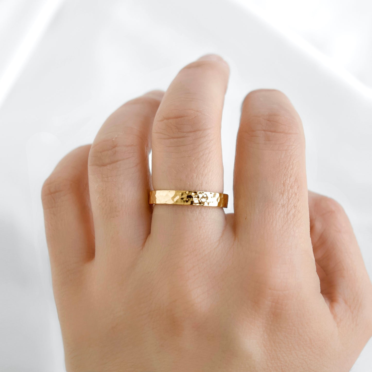 A close-up of a hand wearing a thin, hammered gold band ring against a bright white background.