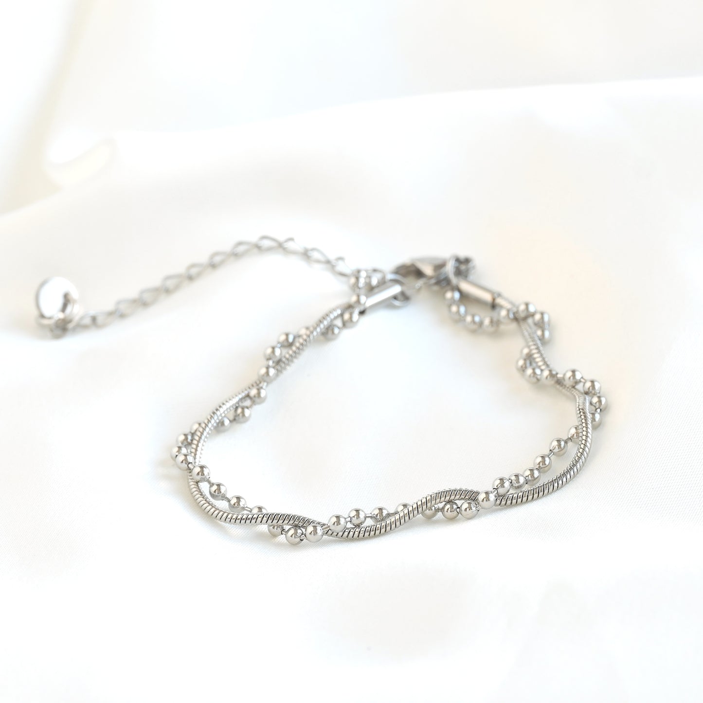 A silver-toned beaded snake chain bracelet with a textured snake-style center band and an adjustable extender chain, displayed on a soft white fabric background.
