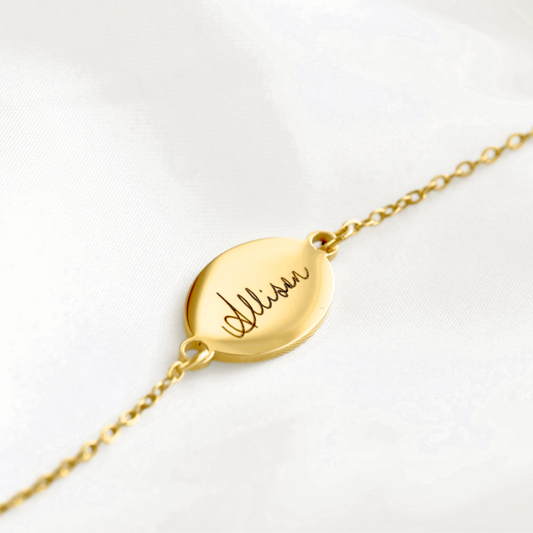 A gold chain bracelet with a flat oval disc pendant engraved with the name 'Allison' in cursive, displayed on a soft white fabric background.