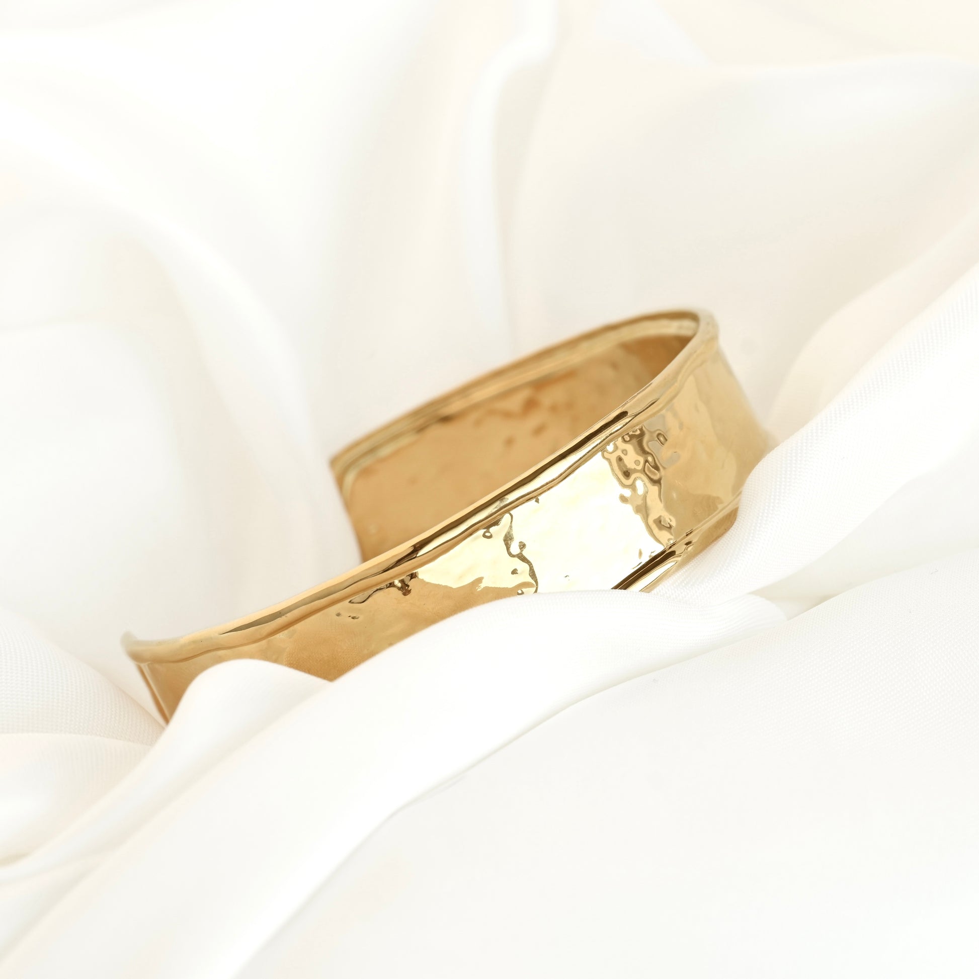 A polished gold cuff bracelet with a hammered texture, resting on draped white satin fabric.