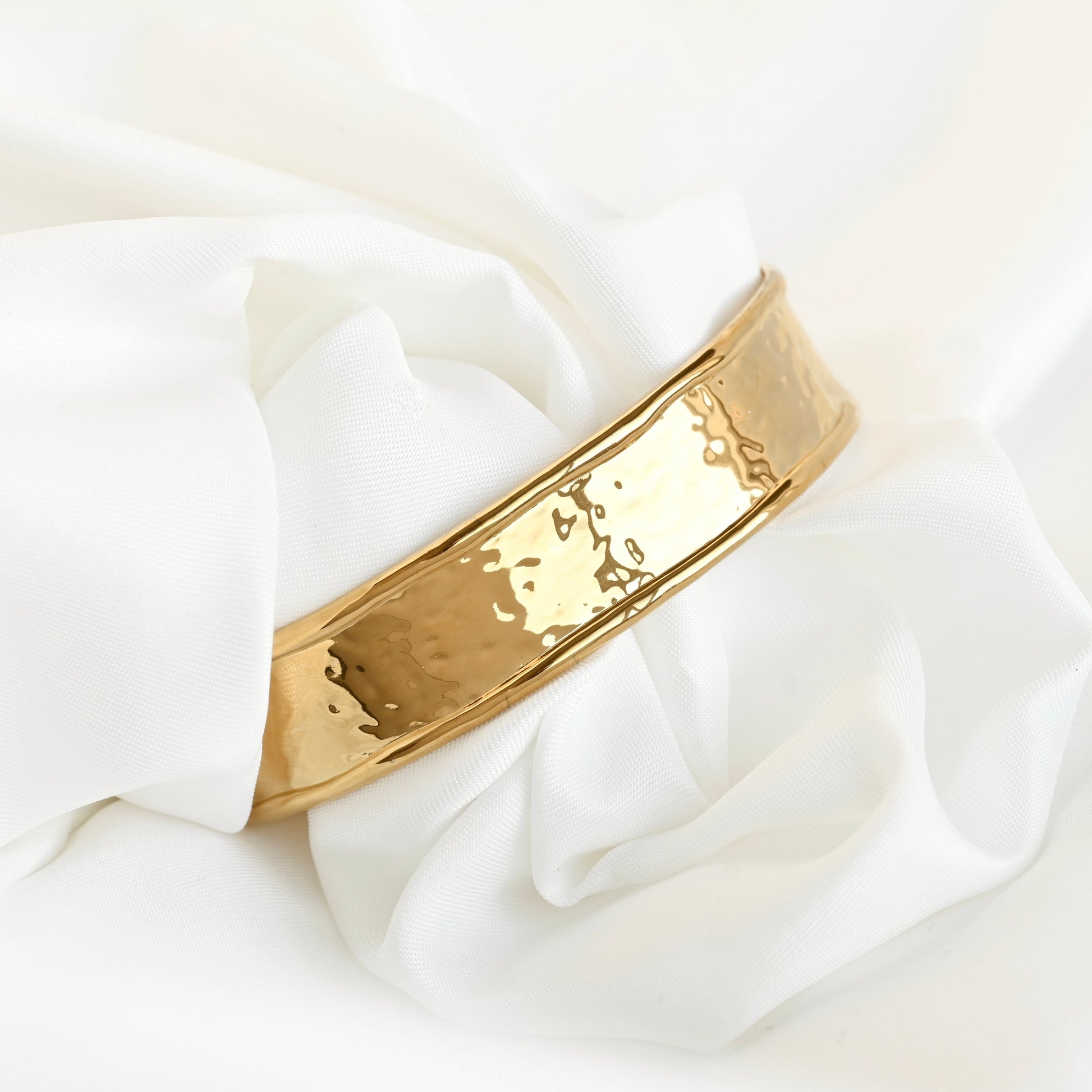 A wide gold-toned metal cuff bracelet with a hammered texture, resting on draped white fabric.